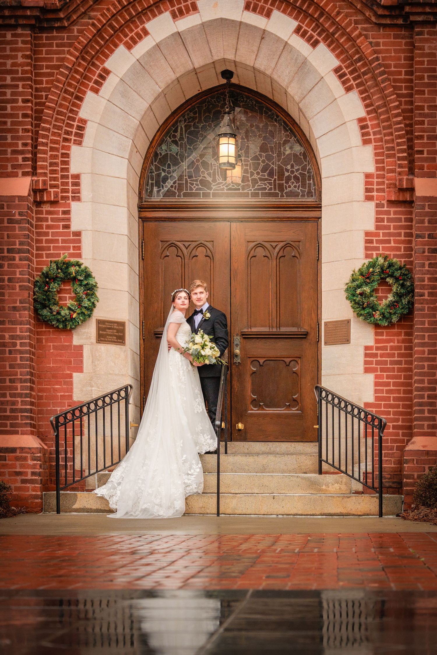 A wedding couple stands in front of an elegant red brick church entrance with arched doorways and hanging floral baskets.