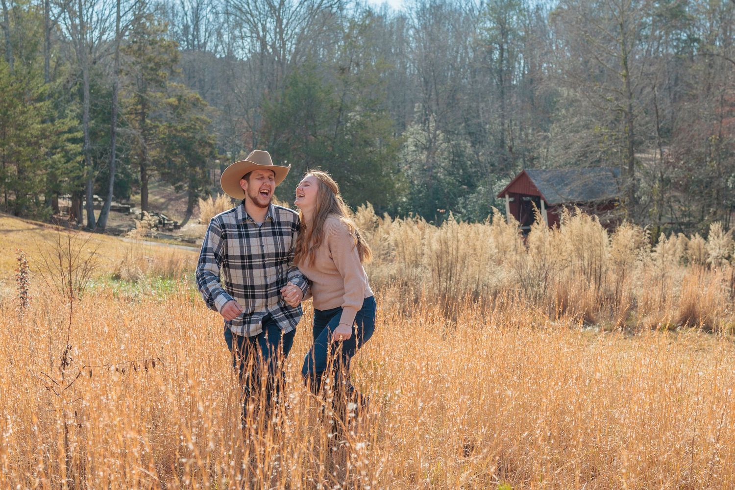 A couple walks together through a golden autumn field with tall grass and trees in the background.