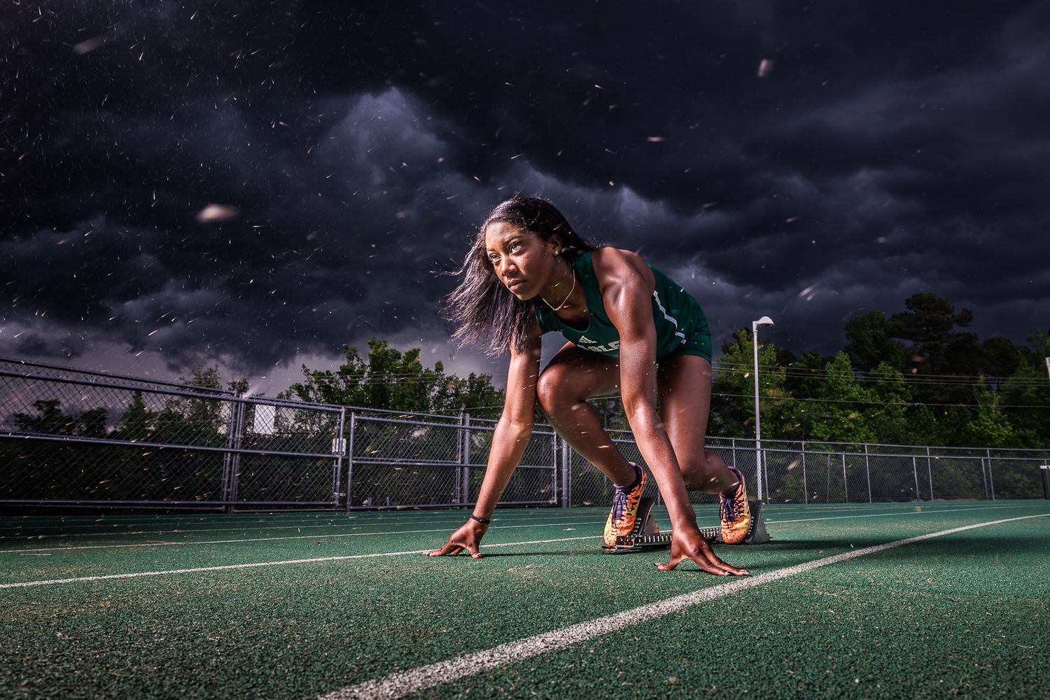 Runner in athletic gear crouches in starting position on green turf under dramatic sky.