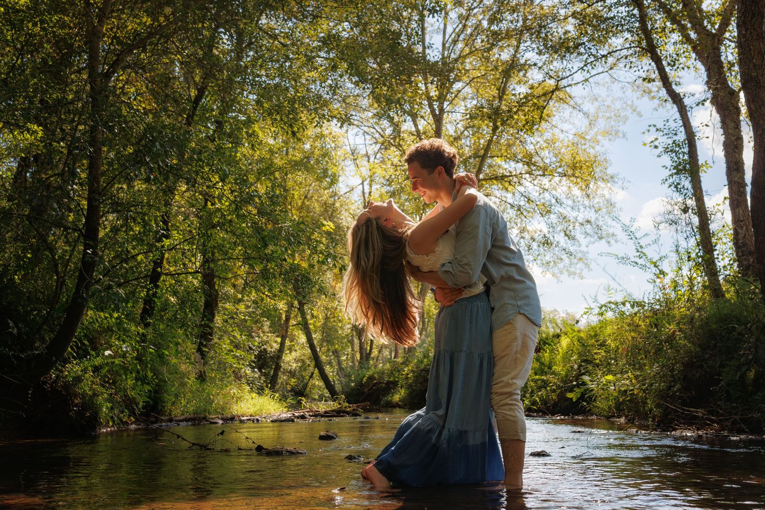 Romantic couple embraces while standing in a sunlit stream surrounded by lush green trees on a summer day.