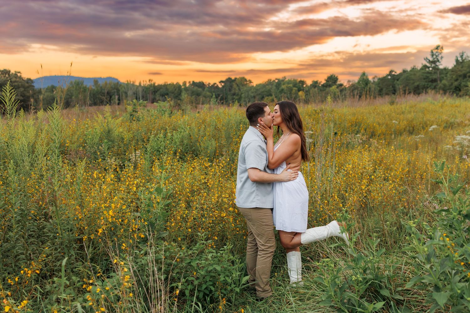 A couple embraces in a golden wildflower meadow at sunset with dramatic orange and pink clouds overhead.