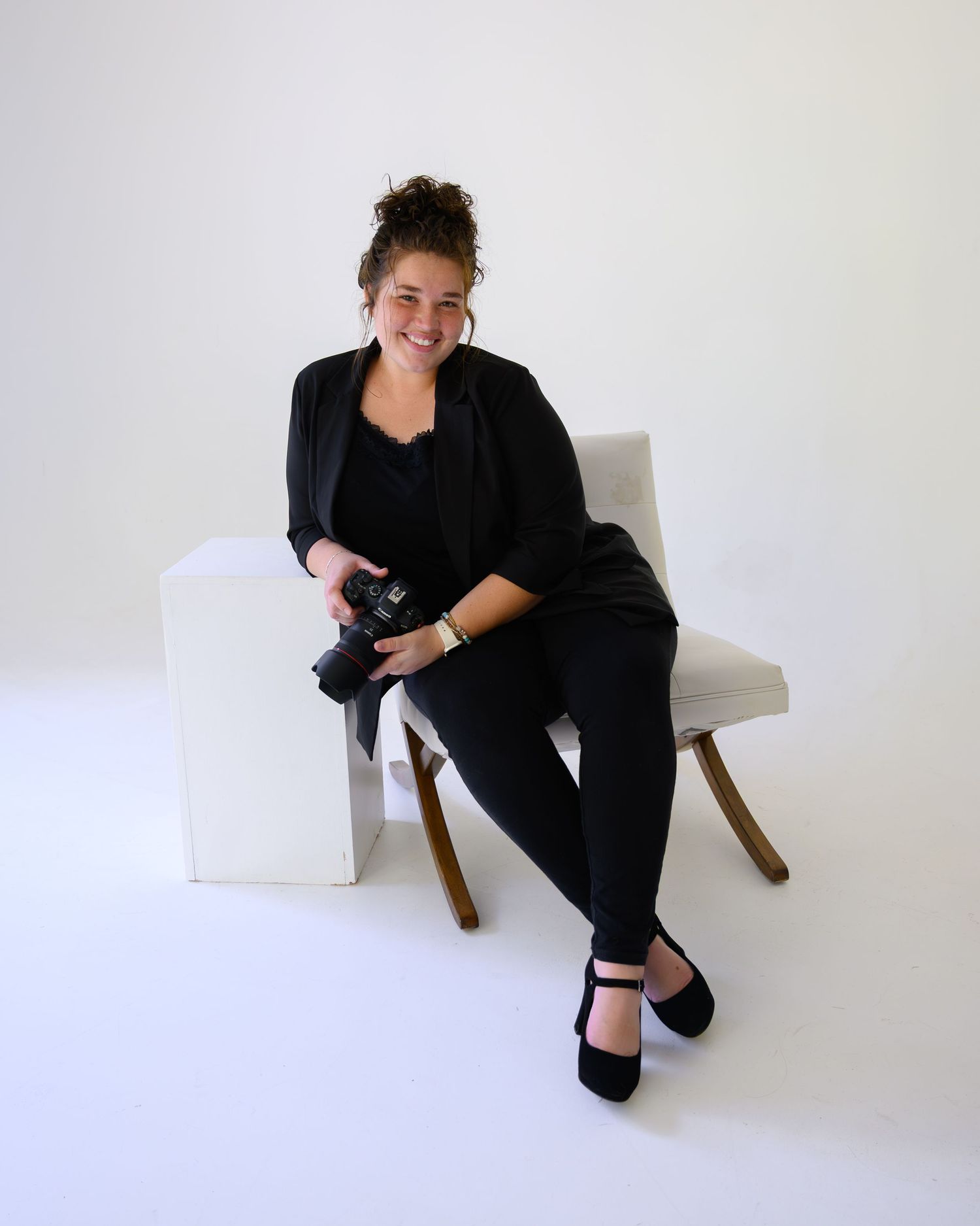 Person in black attire seated on wooden chair against white wall in artistic photo series.