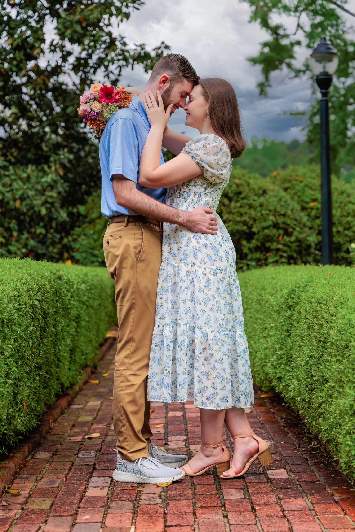 A romantic couple shares intimate moments on a brick pathway surrounded by lush green hedges in a garden setting.