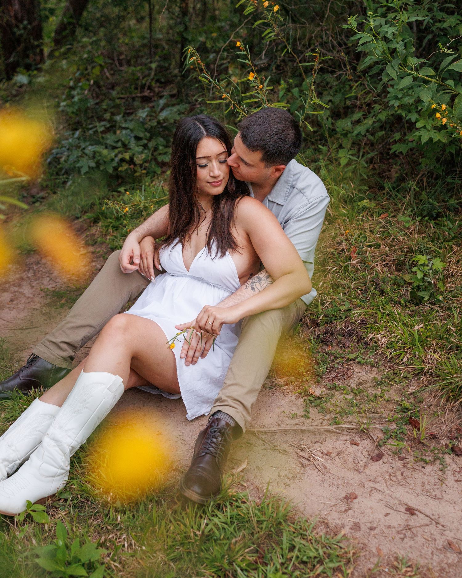 Romantic couple in white outfits sitting close together on a natural path with yellow flowers in foreground.