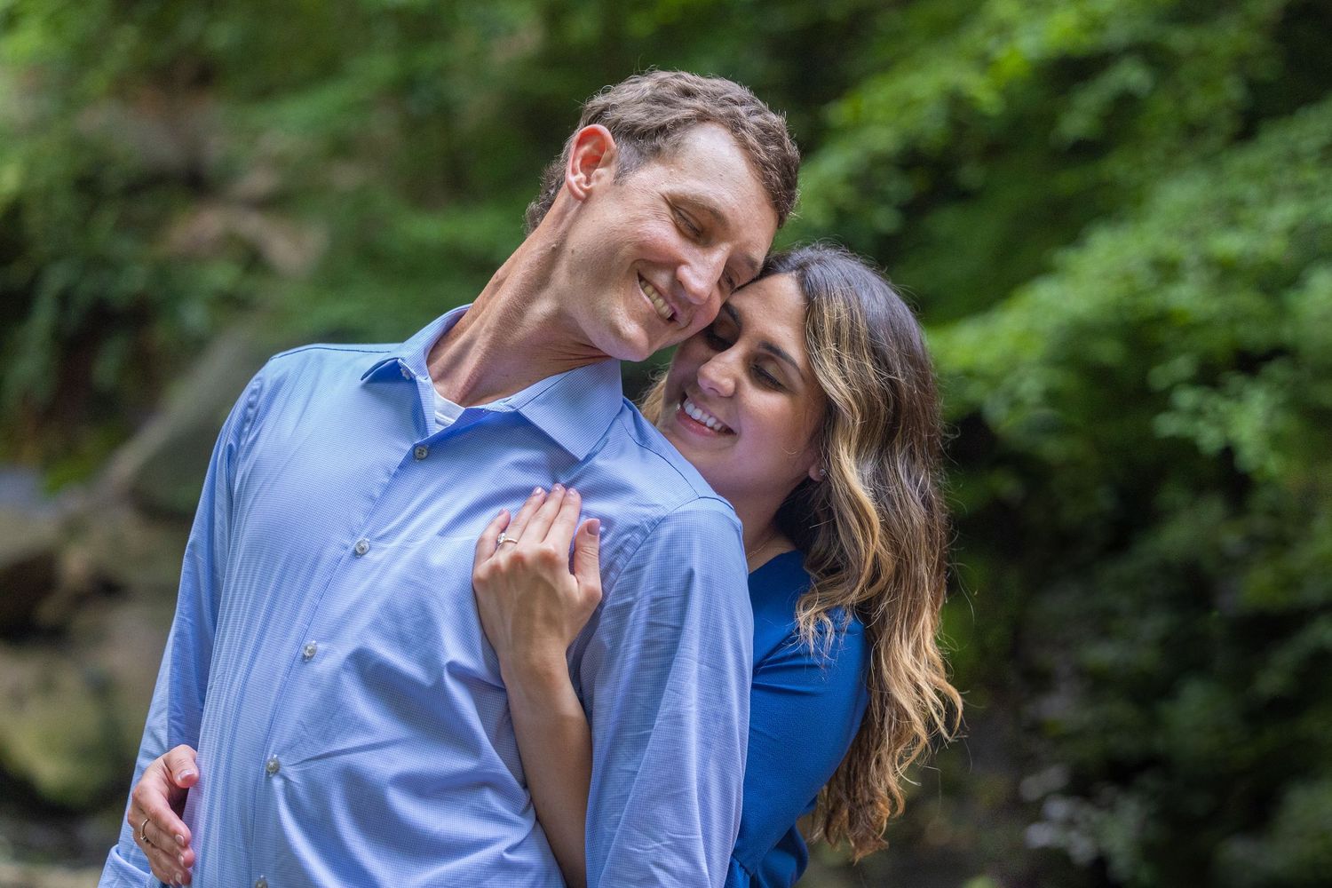 A couple in coordinating blue outfits share an intimate embrace while standing outdoors surrounded by greenery.