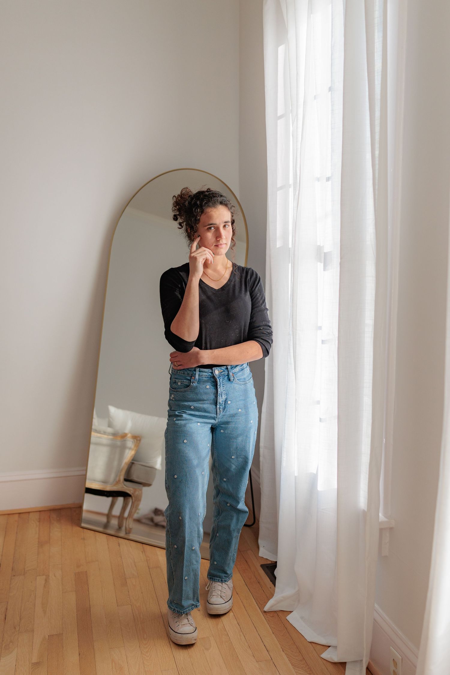 Person in denim overalls and glasses poses playfully against white curtains in a room with hardwood floors.