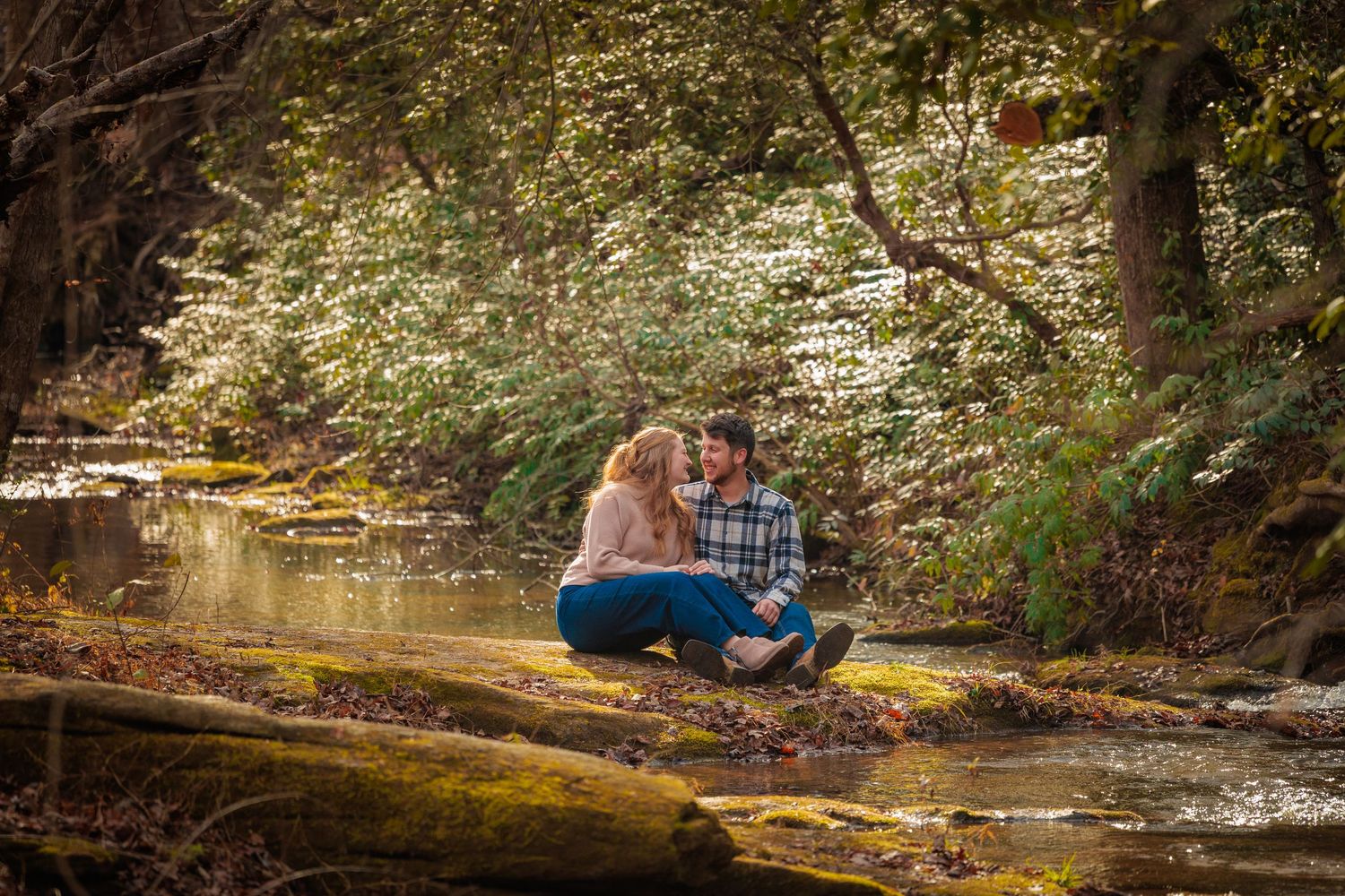 A couple sits together on a mossy log by a flowing creek in a serene autumn forest setting.