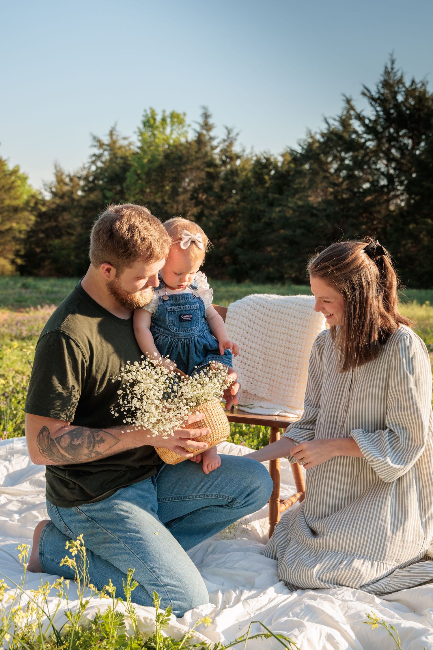 Family having outdoor picnic on white blanket with baby's breath flowers in sunset light near evergreen trees.