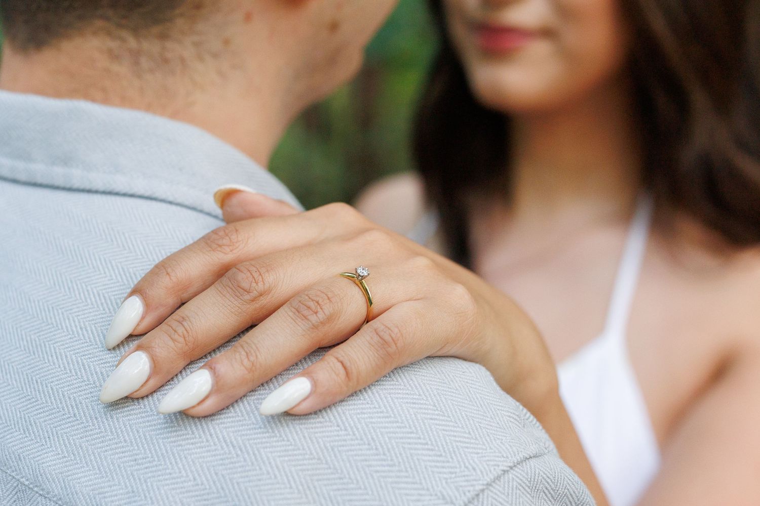 Close-up view of an engagement ring on a hand with white nail polish against a light background.