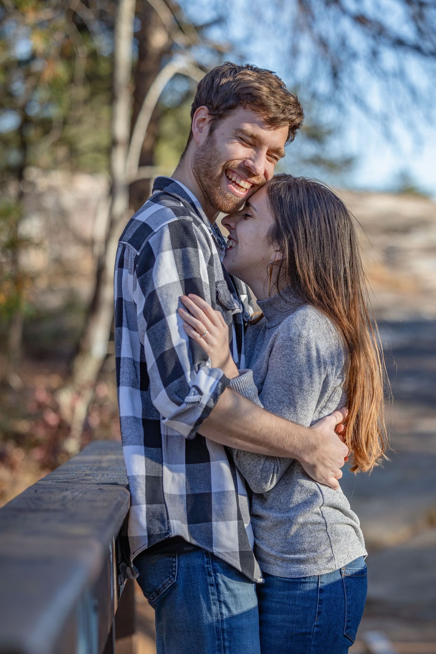 A couple embraces and smiles while standing together outside on a sunny day wearing casual clothing.