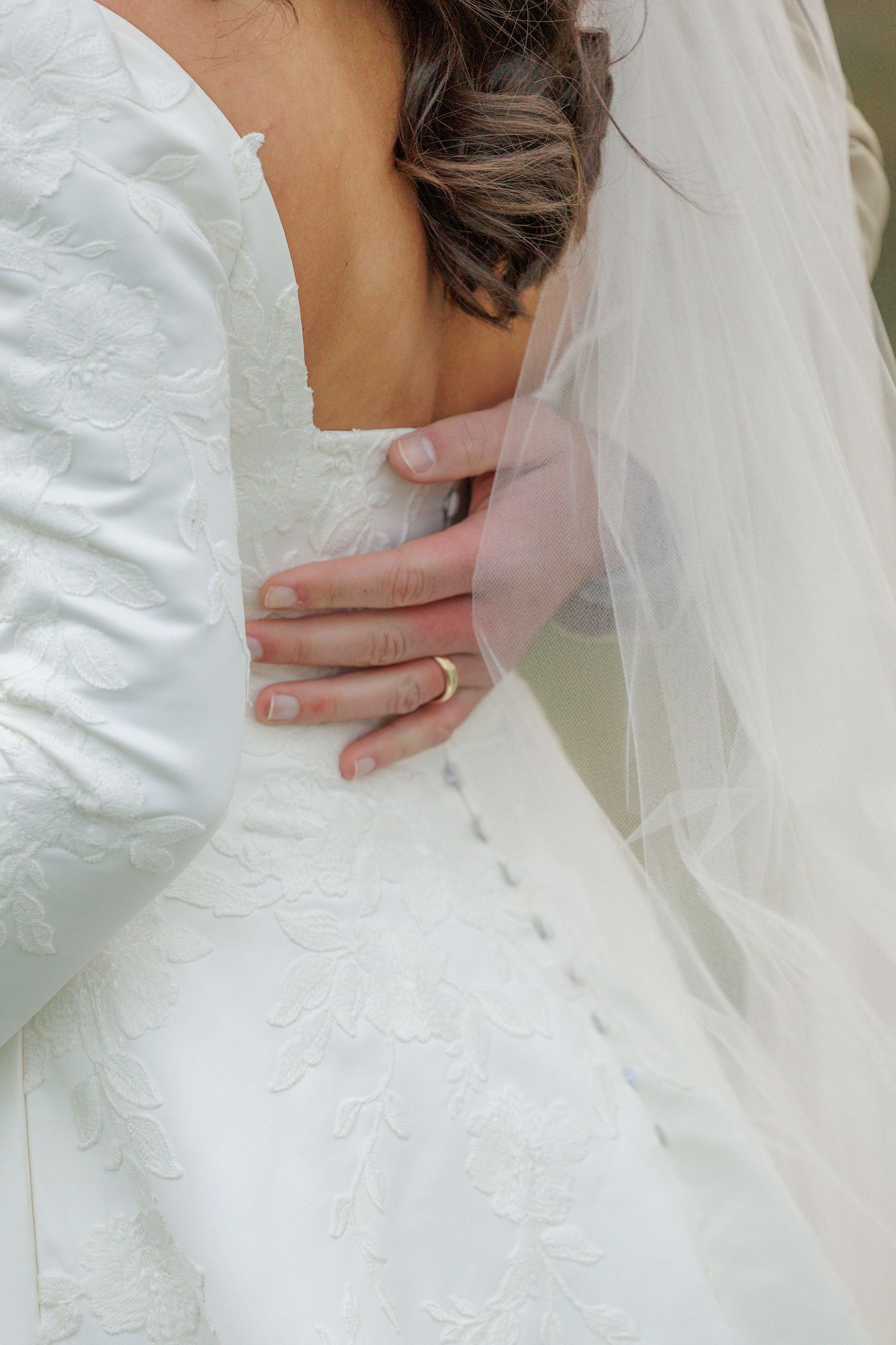 A hand tenderly touches the waist of a white wedding dress with delicate lace detailing and flowing veil.