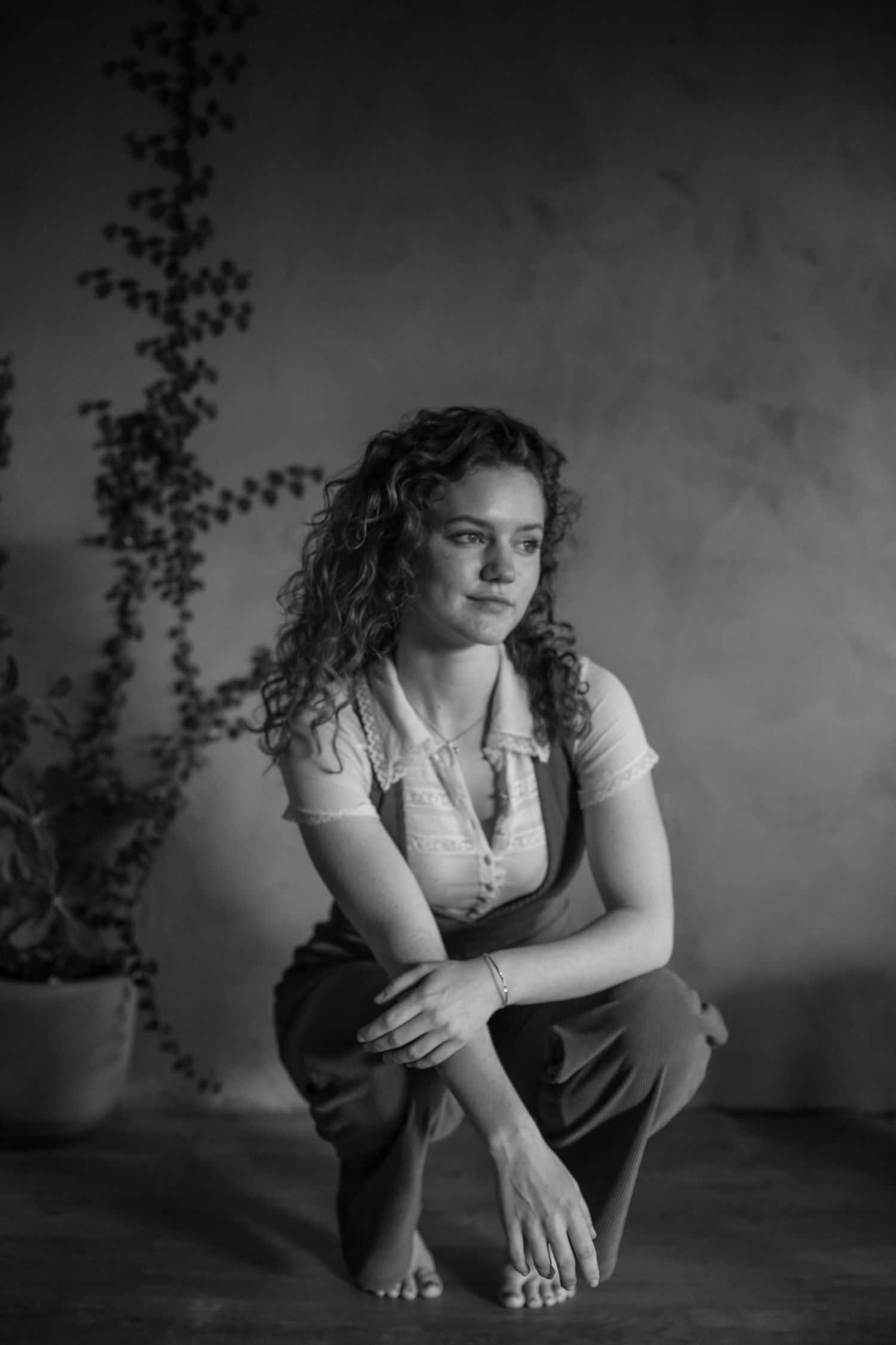 Black and white portrait of a woman with curly hair crouching barefoot, wearing casual attire in a moody setting.
