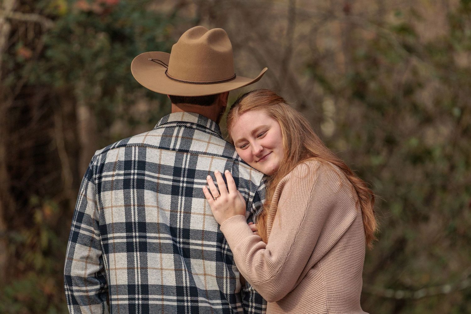 Romantic couple in western attire embracing outdoors with a cowboy hat and plaid shirt.