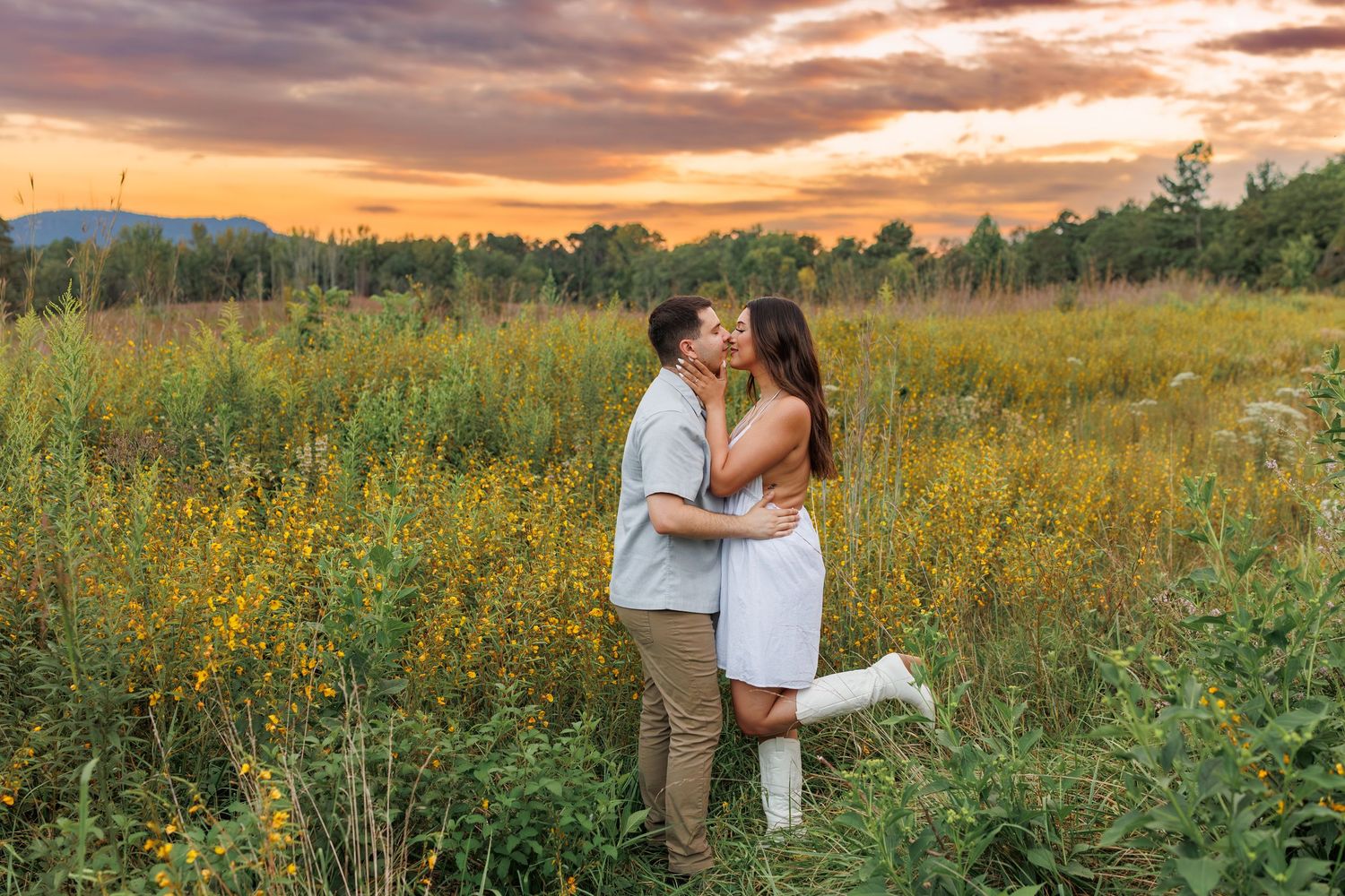 A couple embraces in a golden wildflower field at sunset with dramatic orange and purple clouds in the sky.