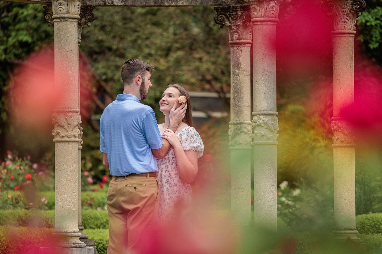 A romantic couple embraces under stone columns in a garden with pink flowers blurring the foreground.