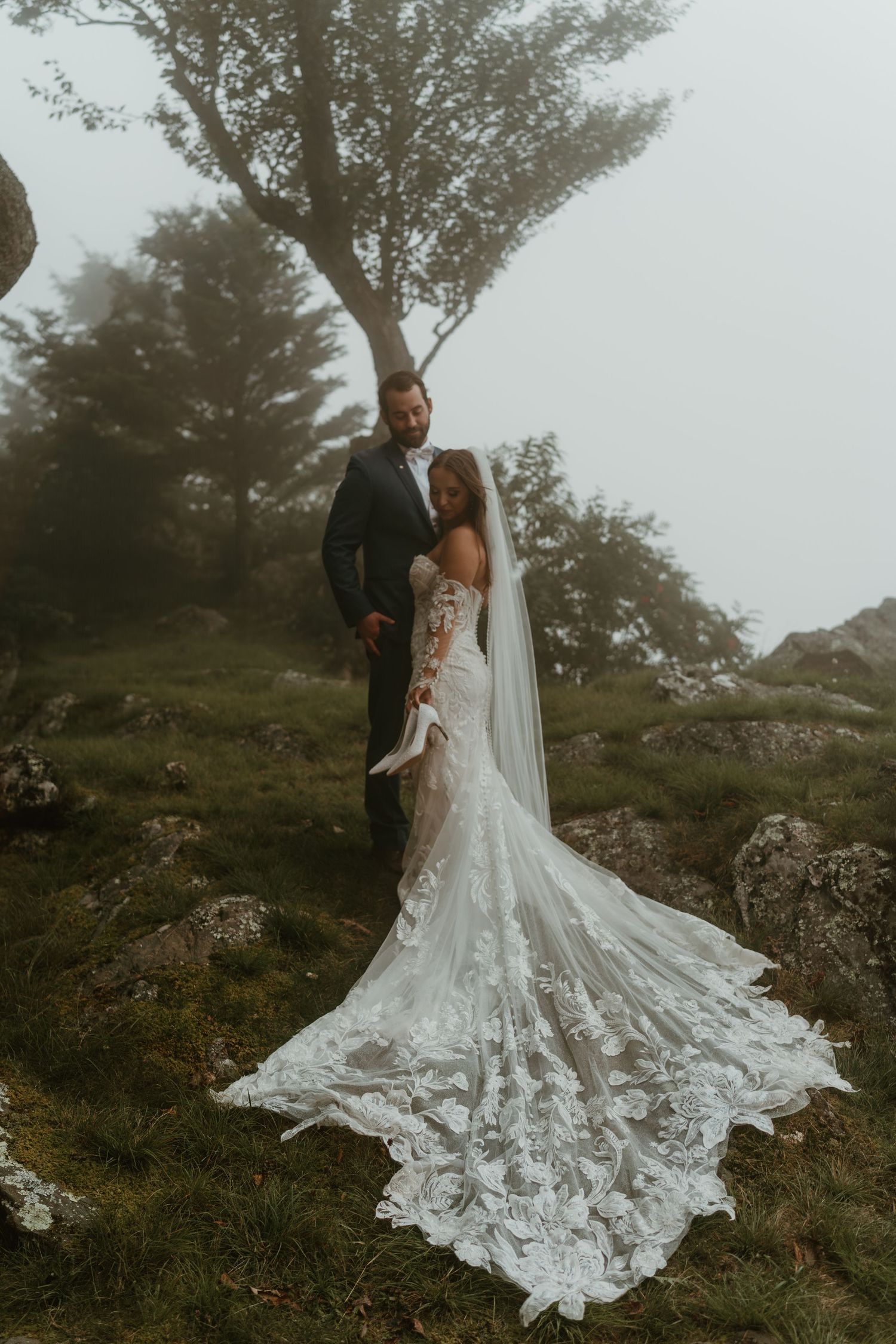 Bride in lace mermaid gown with cathedral veil and groom in black suit posing on misty rocky hillside.