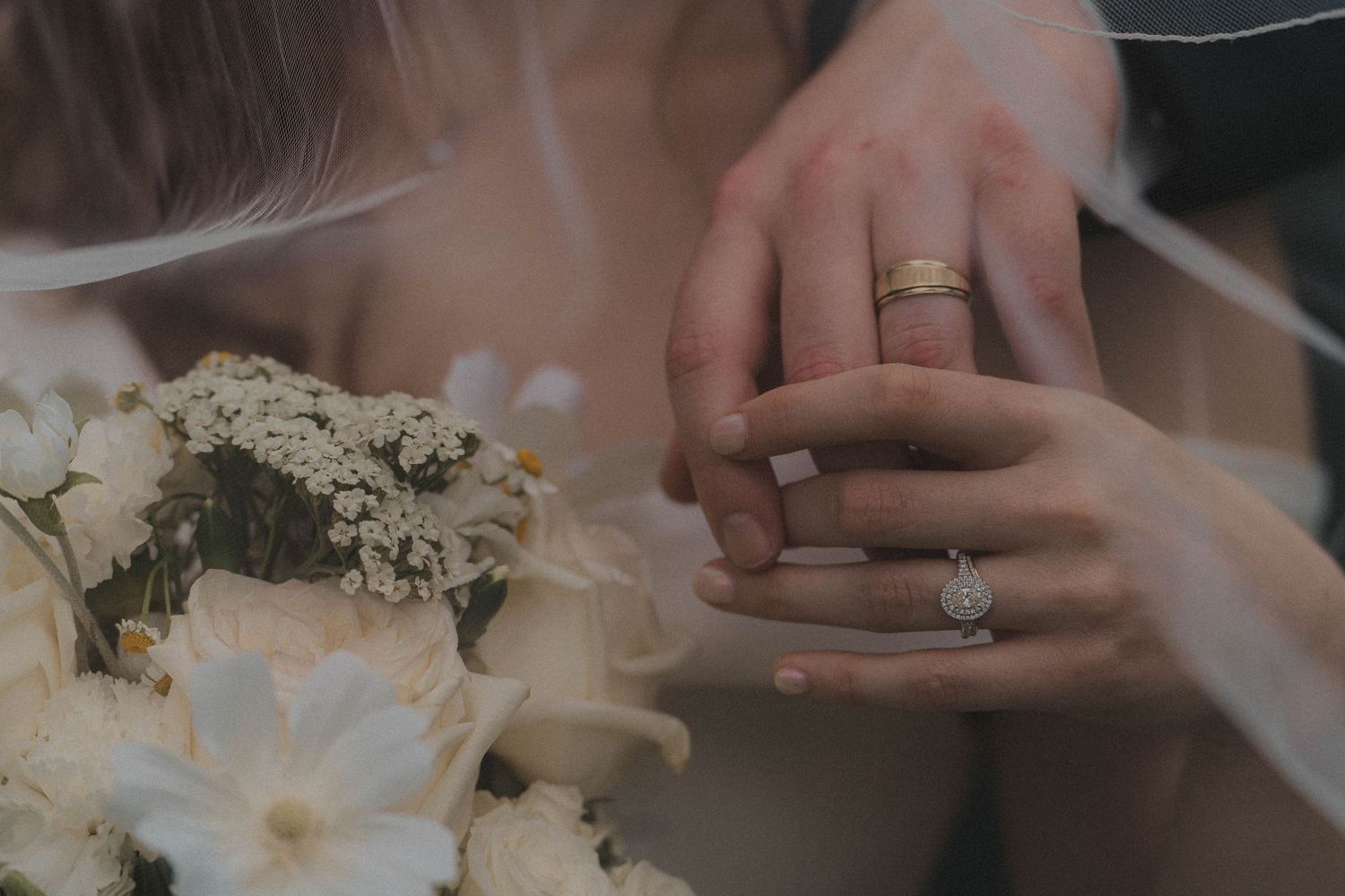 Close-up of bride and groom hands showing wedding rings with white floral bouquet.