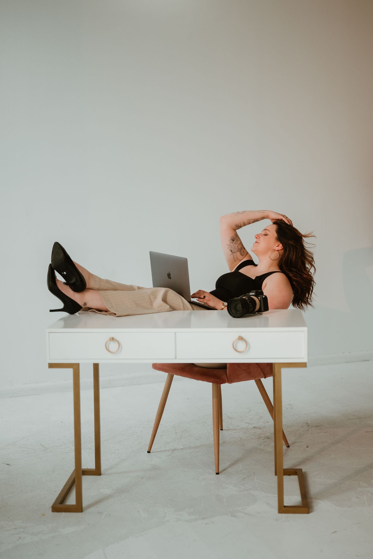 Female photographer relaxing at white gold desk with feet up, laptop and camera, in bright studio space.