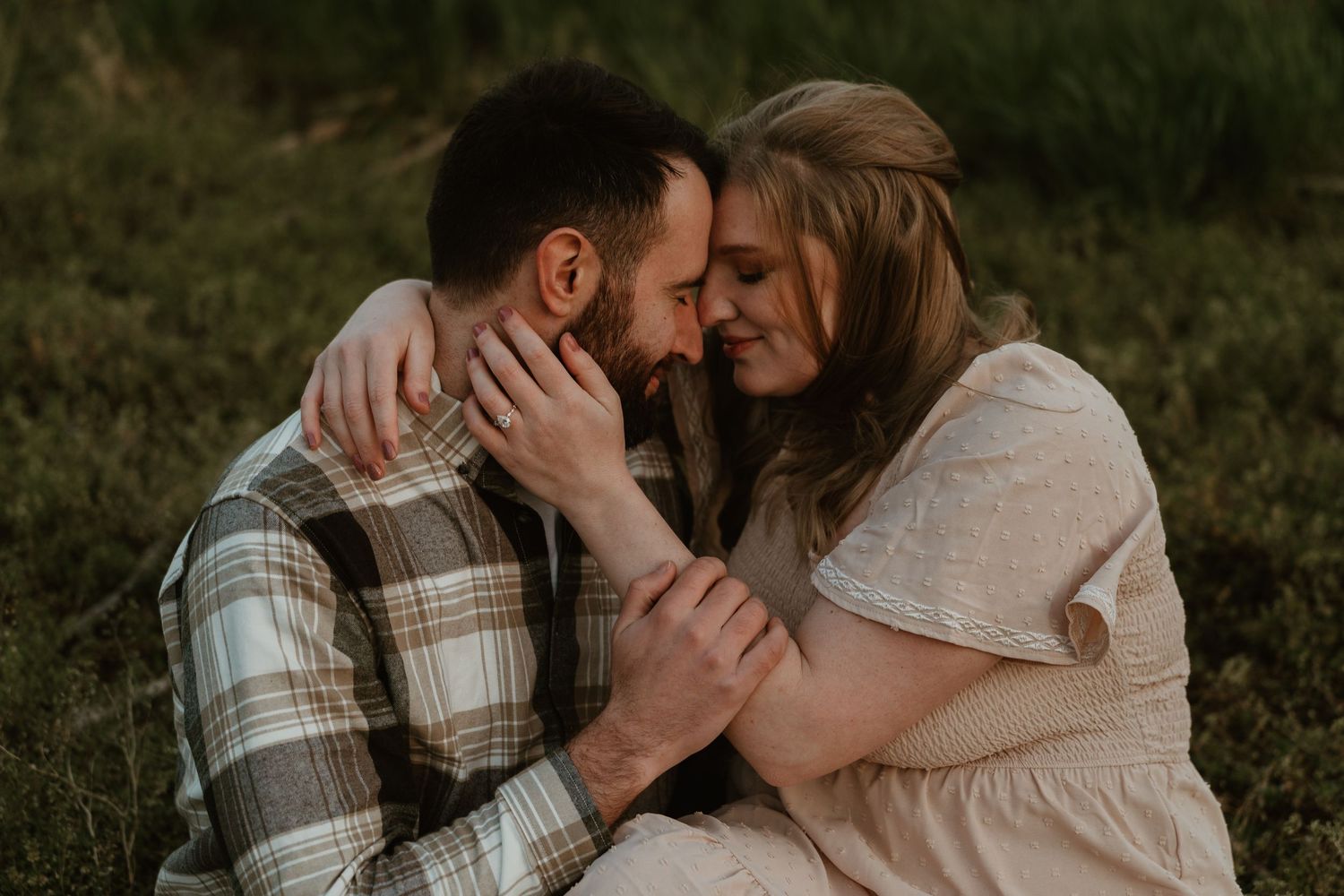 Couple sitting close together in grass, touching foreheads in a tender moment.