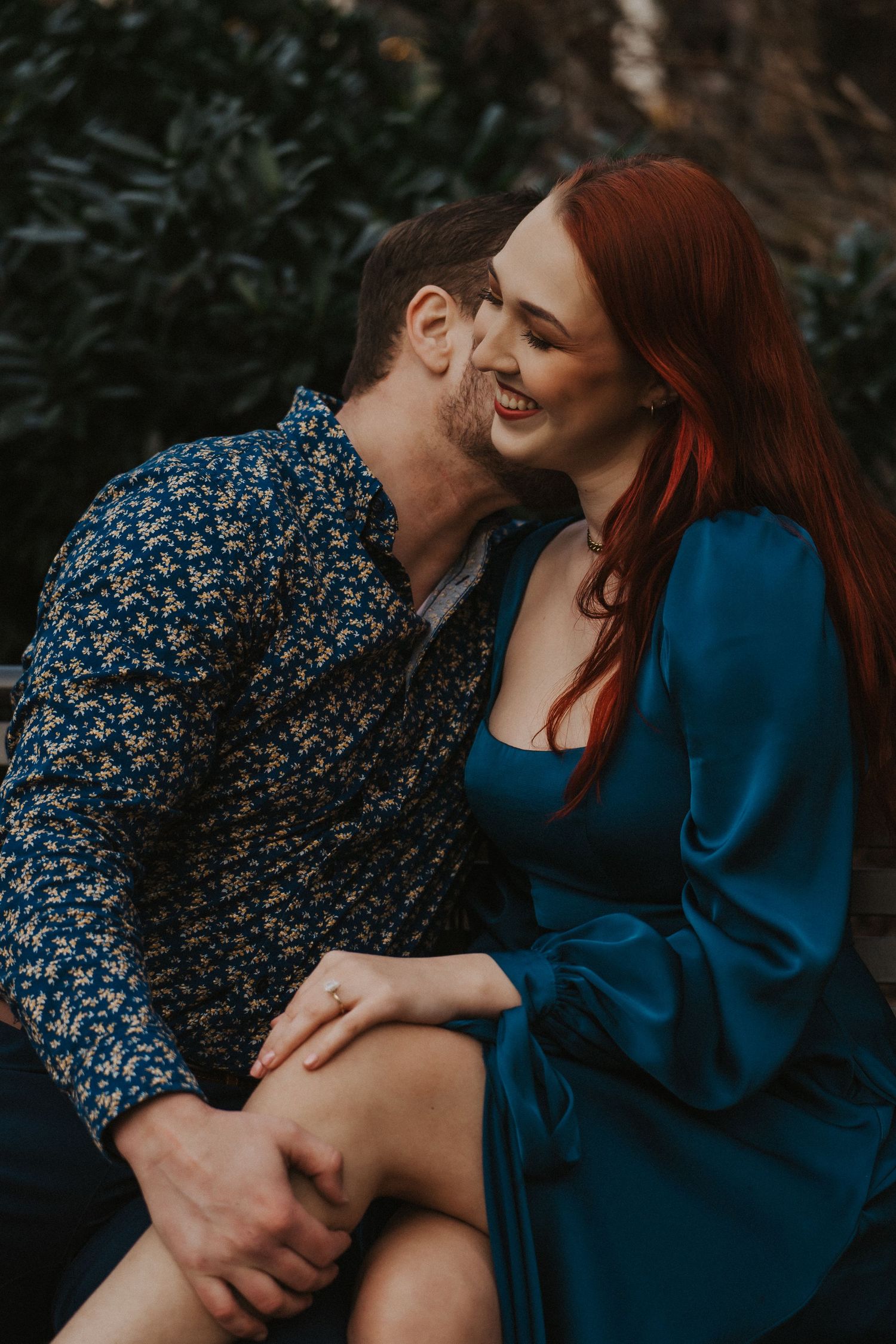 Joyful redhead woman in blue dress laughing as her partner whispers in her ear outdoors.