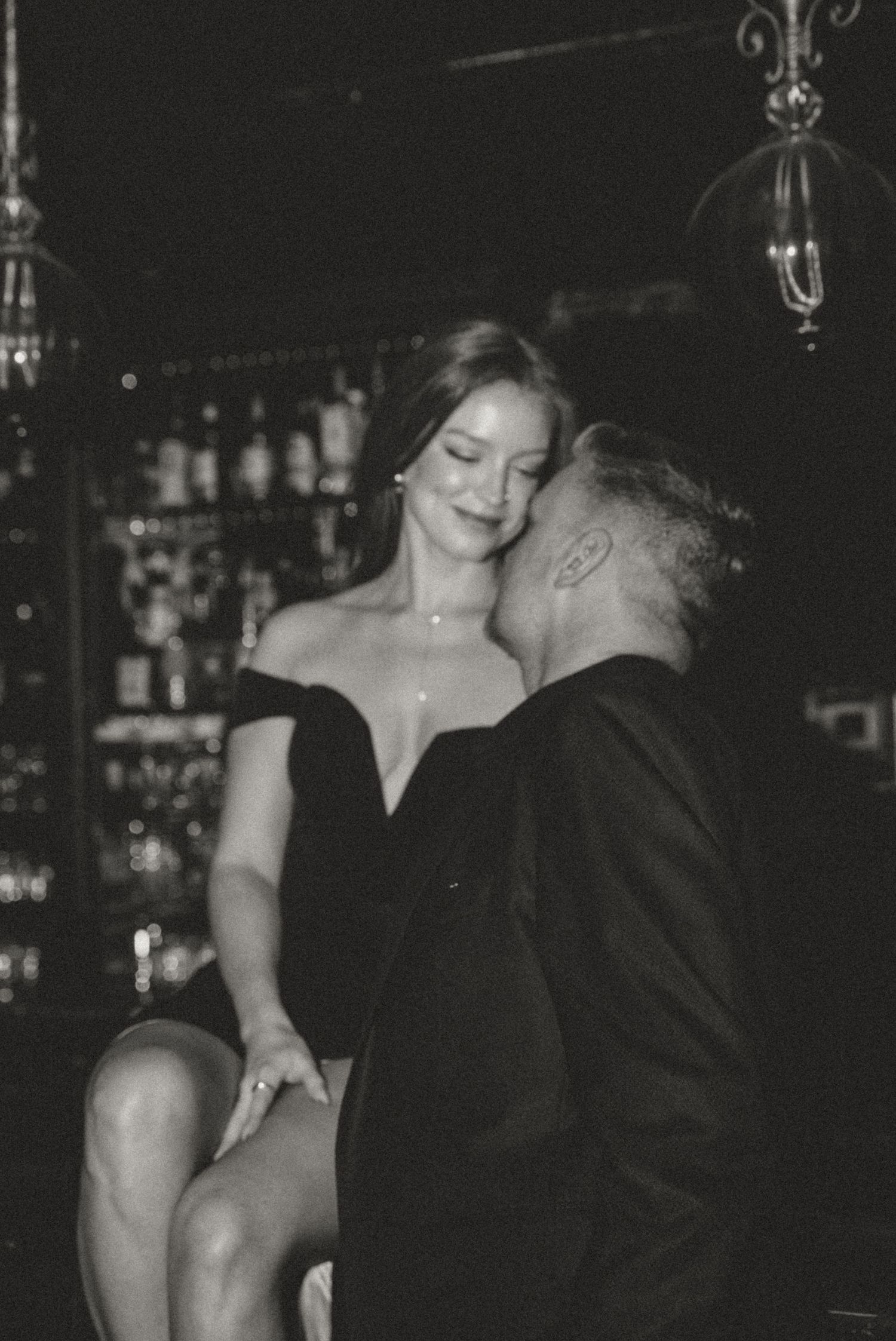Black and white photo of a man kissing a woman's cheek at a dark bar setting.