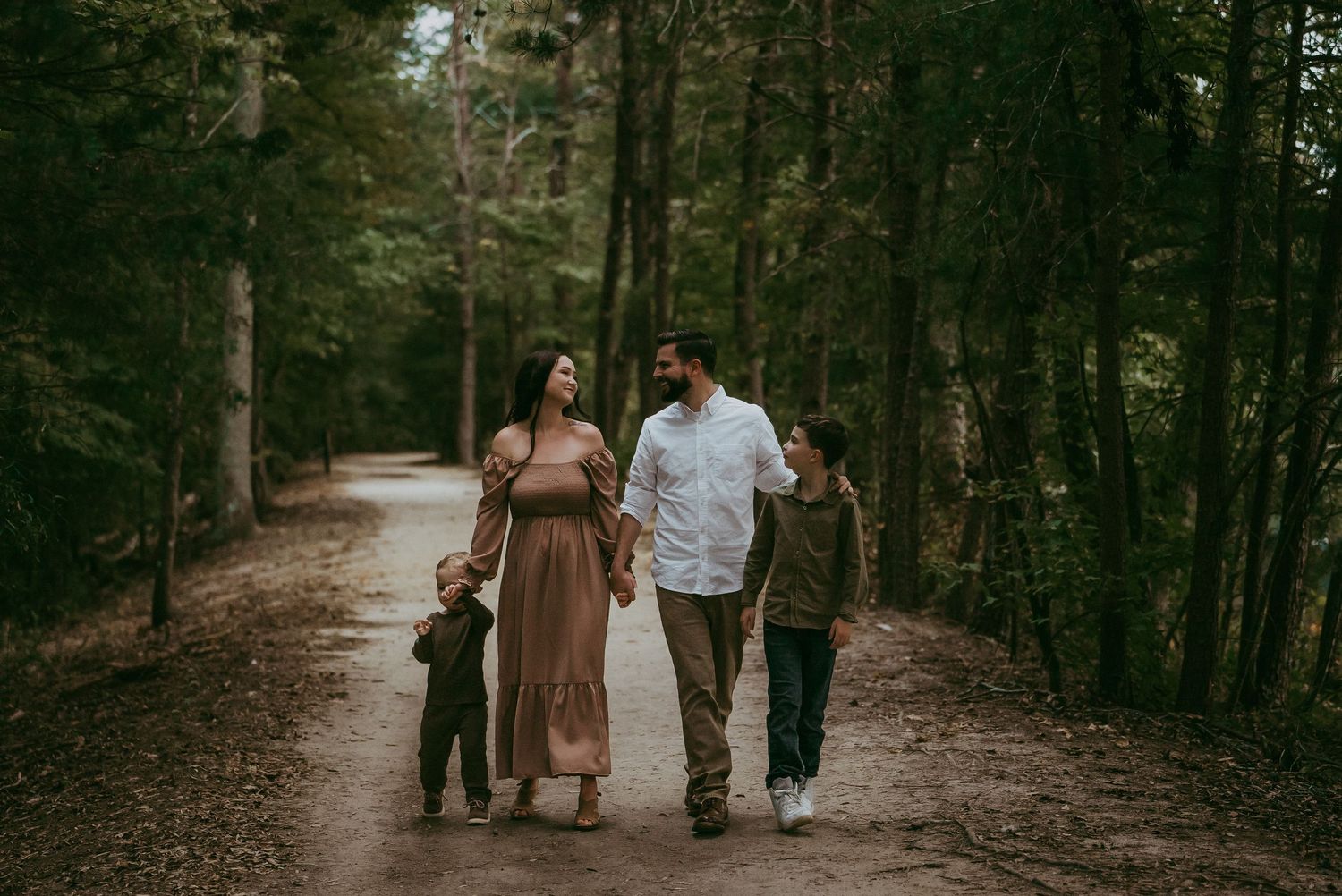 A family of four walks together along a forest path, smiling and holding hands.