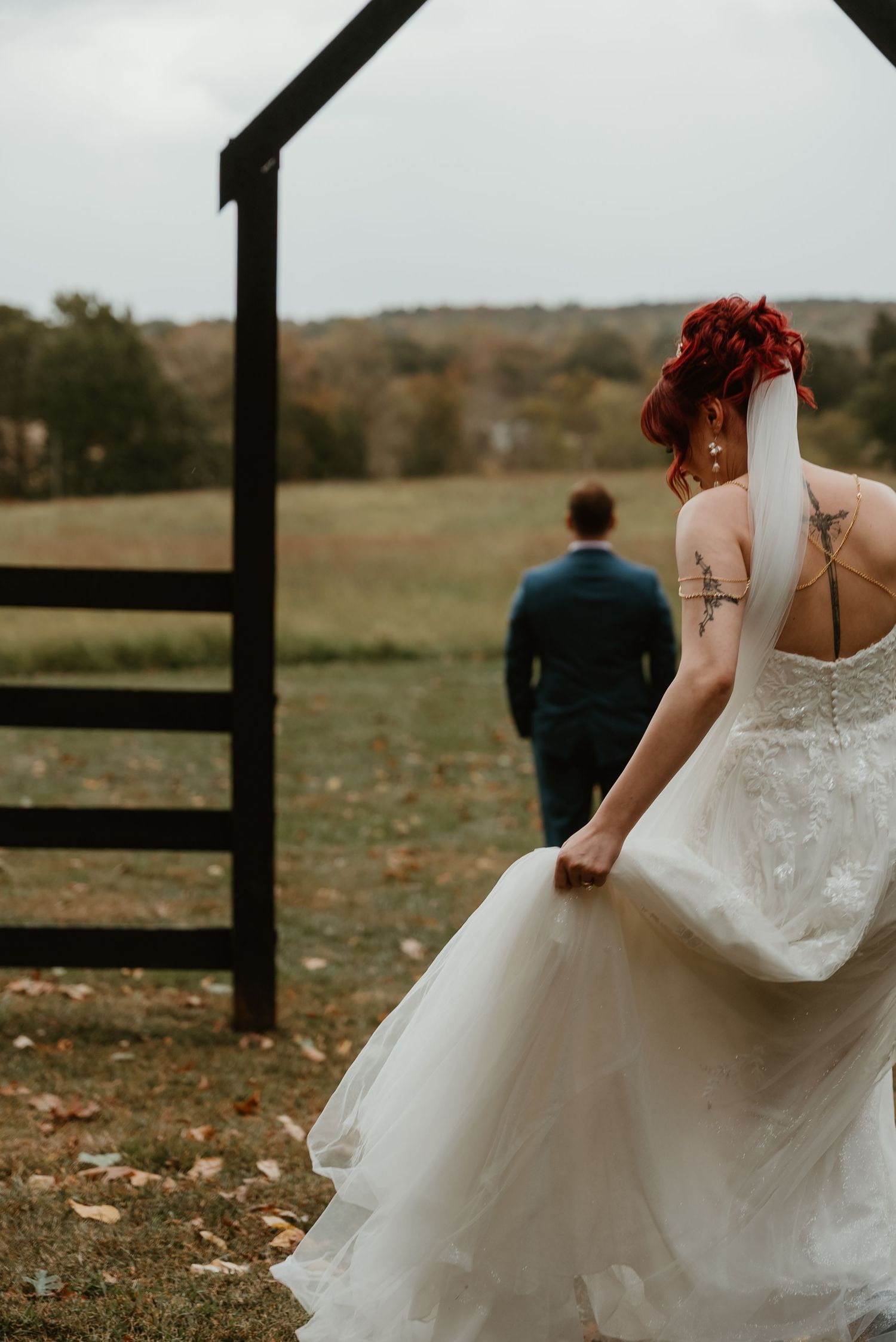 Red-haired tattooed bride in white gown approaching groom standing at rustic wooden arch in autumn field.