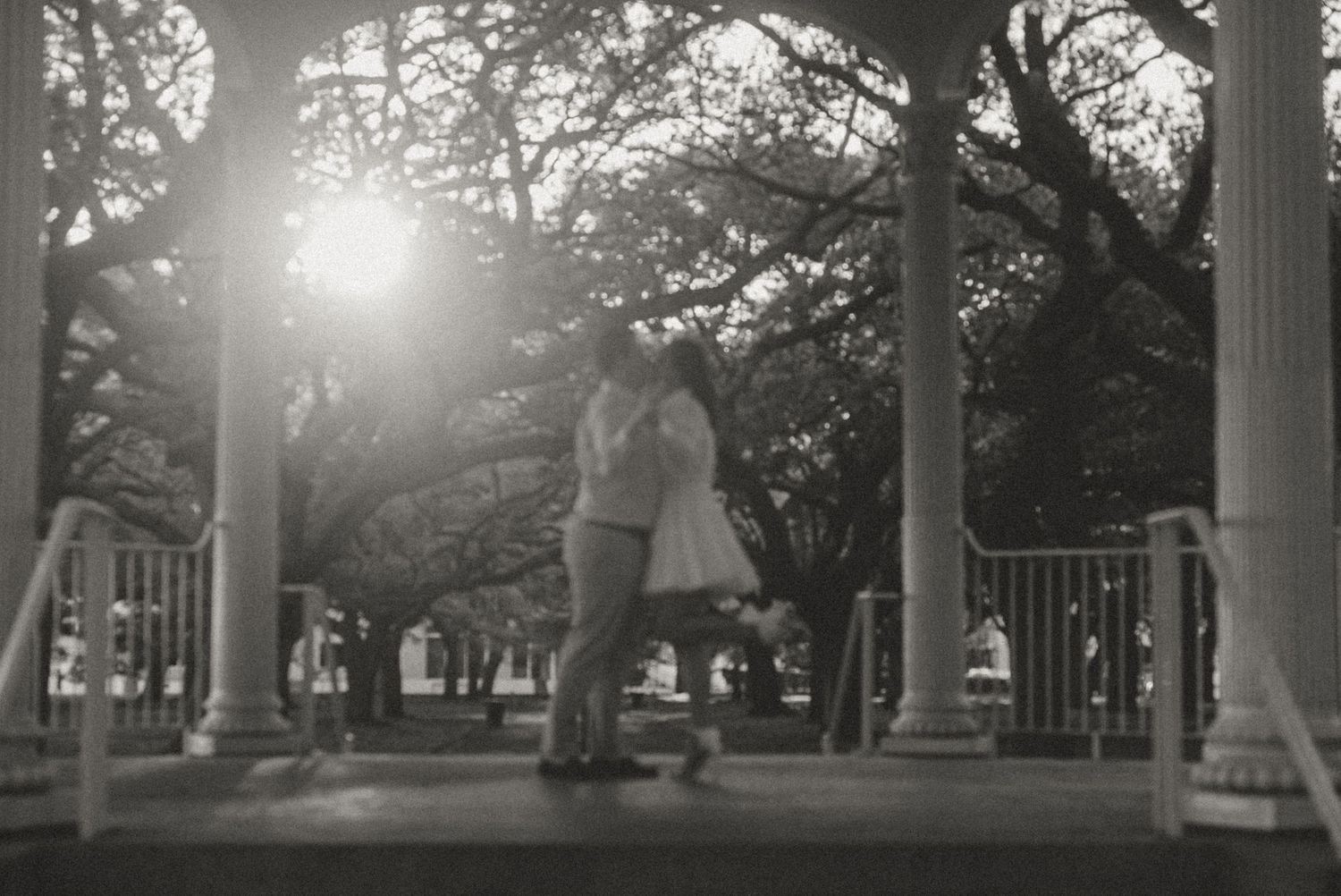 Black and white photo of a couple kissing in a gazebo surrounded by trees with sunlight glowing behind them.