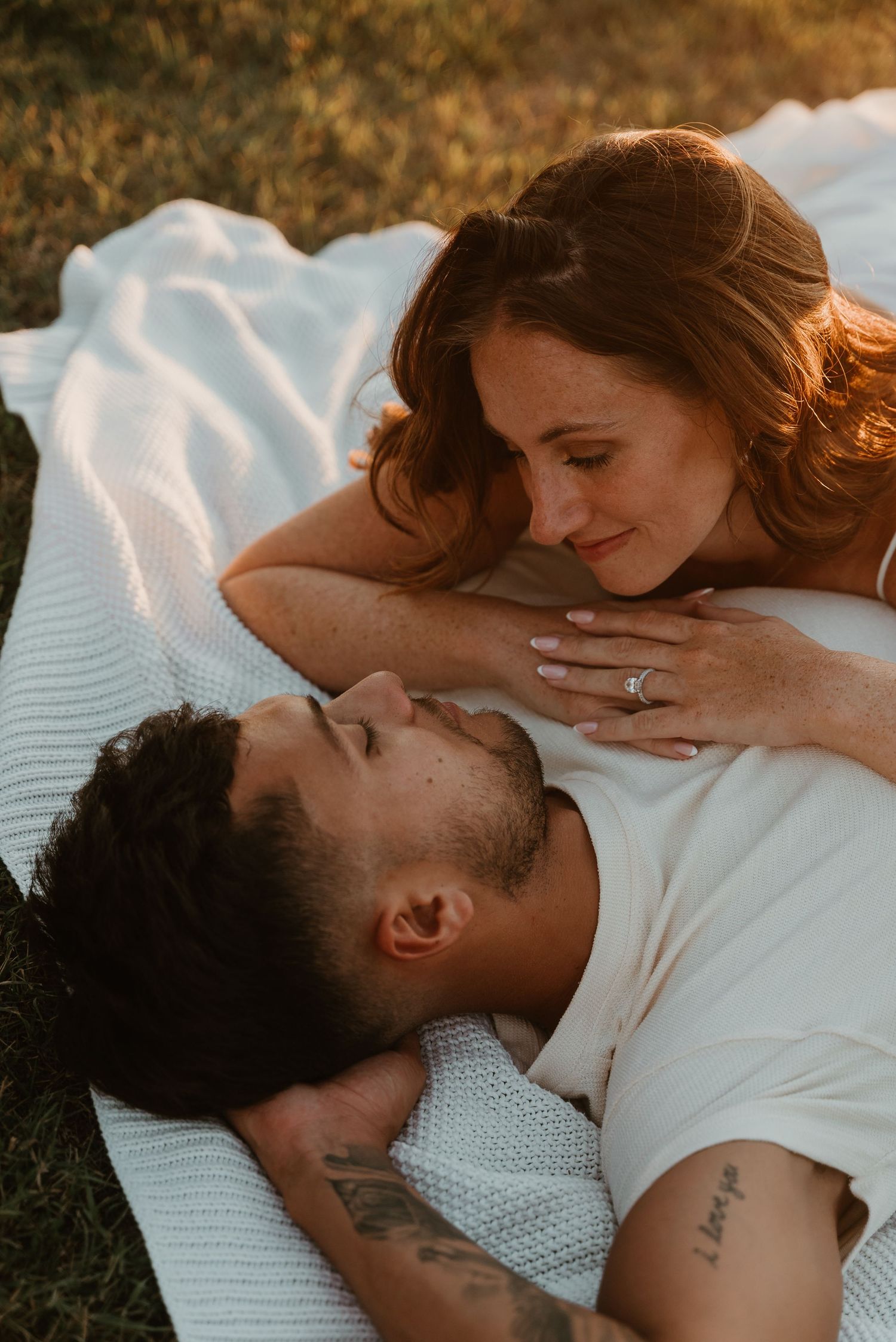 Couple lying together on a white blanket sharing a tender gaze during sunset engagement photos.