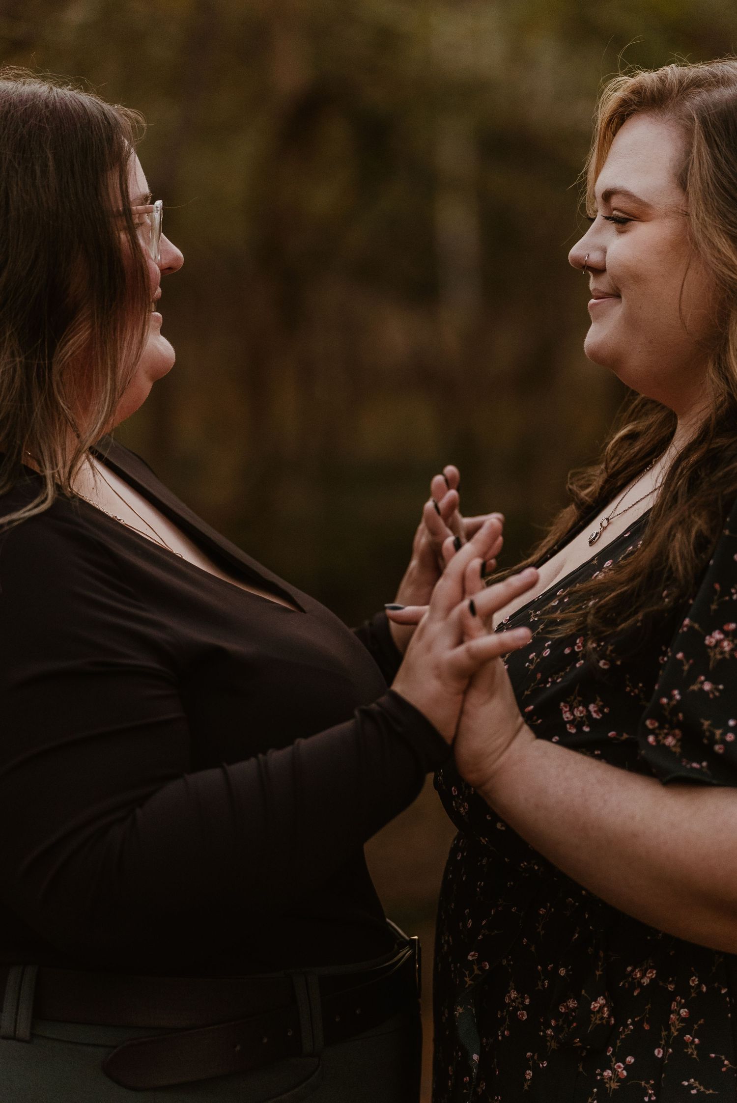Two women holding hands and smiling at each other outdoors in autumn.