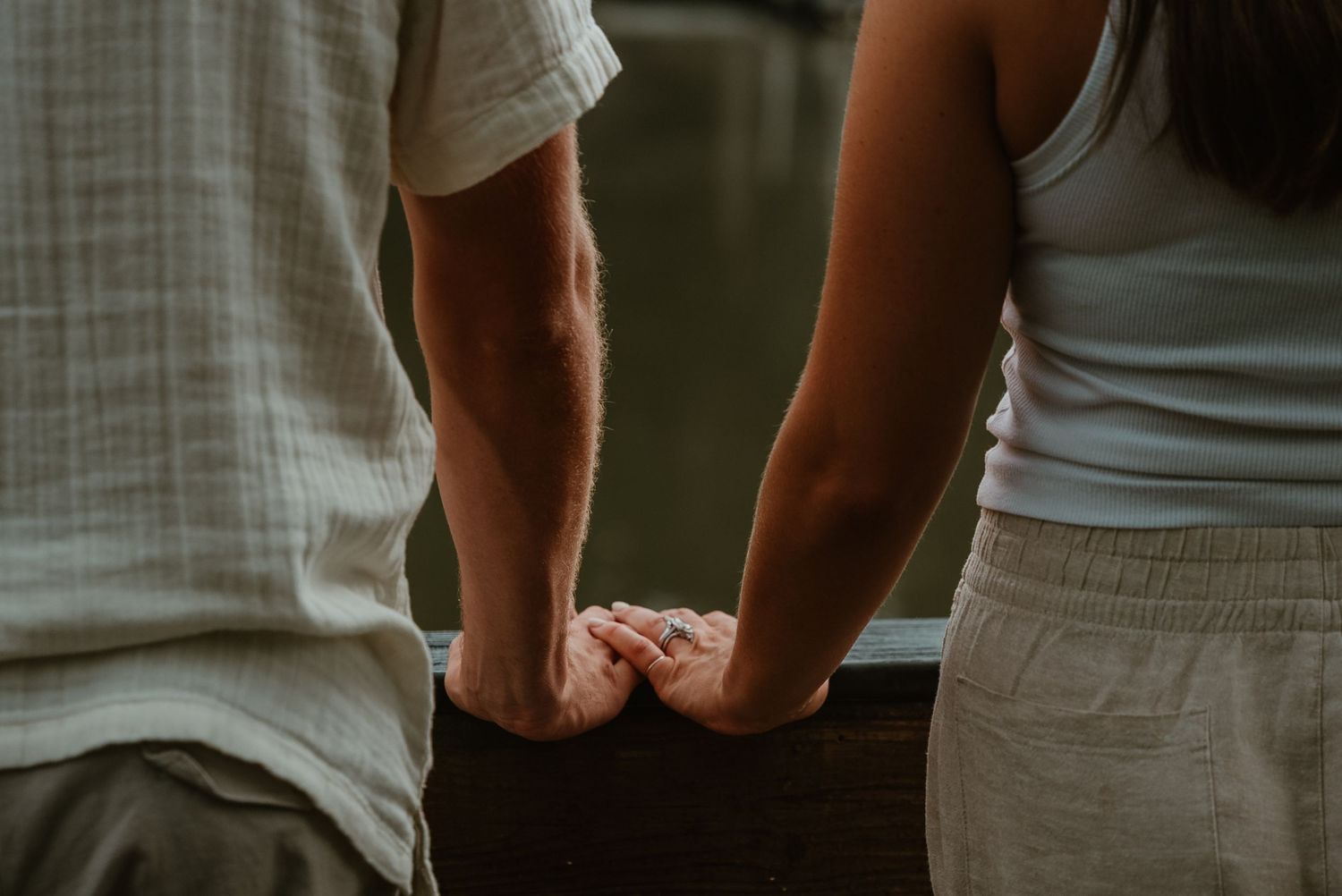 Close-up of a couple holding hands on a wooden railing by a lake during an outdoor engagement session.