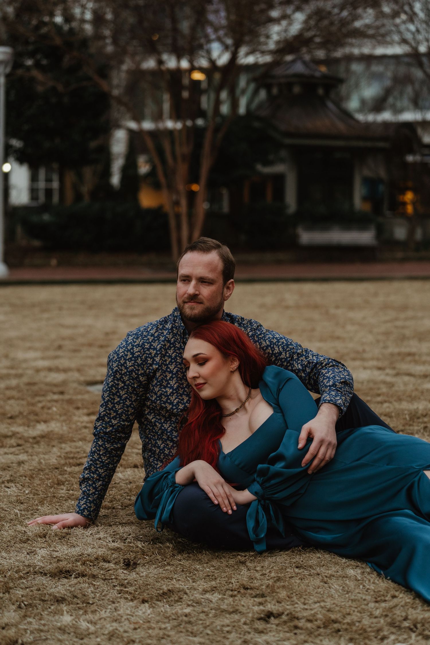 Couple posing on dry grass, woman in teal dress leaning back against man in floral shirt.
