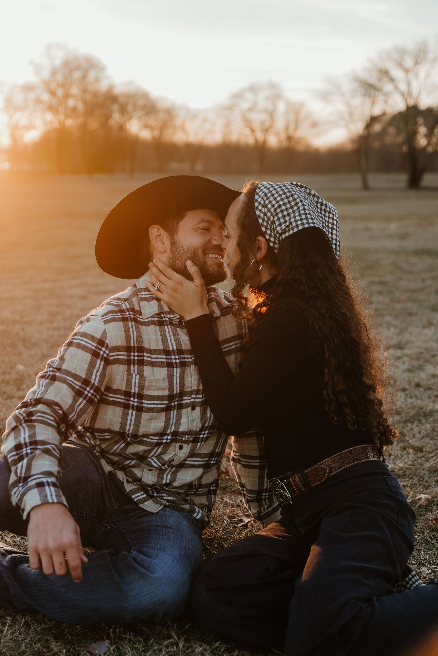 Couple sharing a tender kiss at golden hour sunset in an open field, man wearing a cowboy hat.