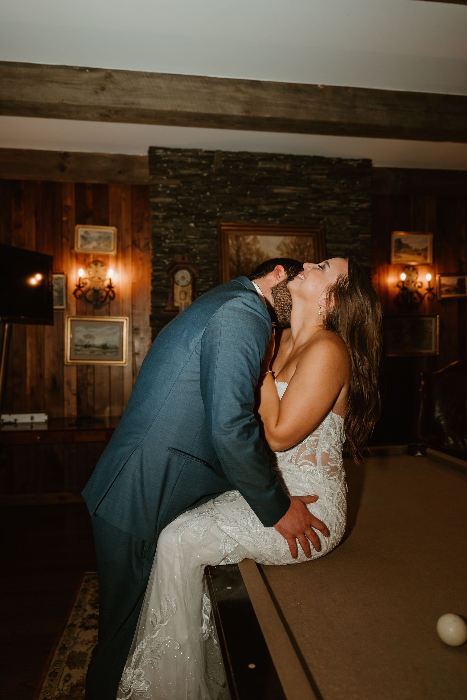 Bride in lace gown laughing as groom dips her over pool table in rustic wood-paneled reception room.