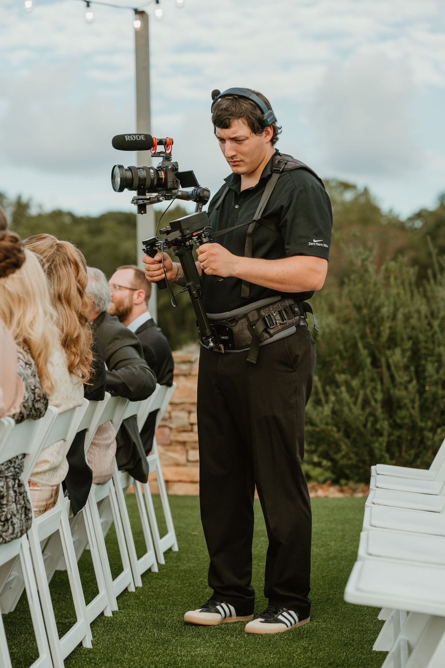 Wedding videographer using stabilized camera rig to film outdoor ceremony with white chairs.