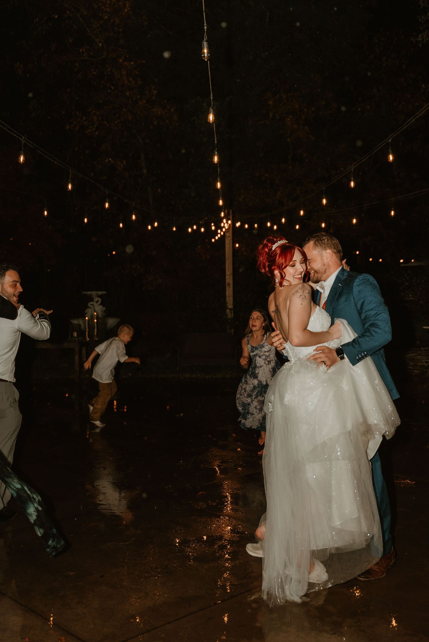 Bride and groom share first dance in the rain under string lights at outdoor wedding reception.