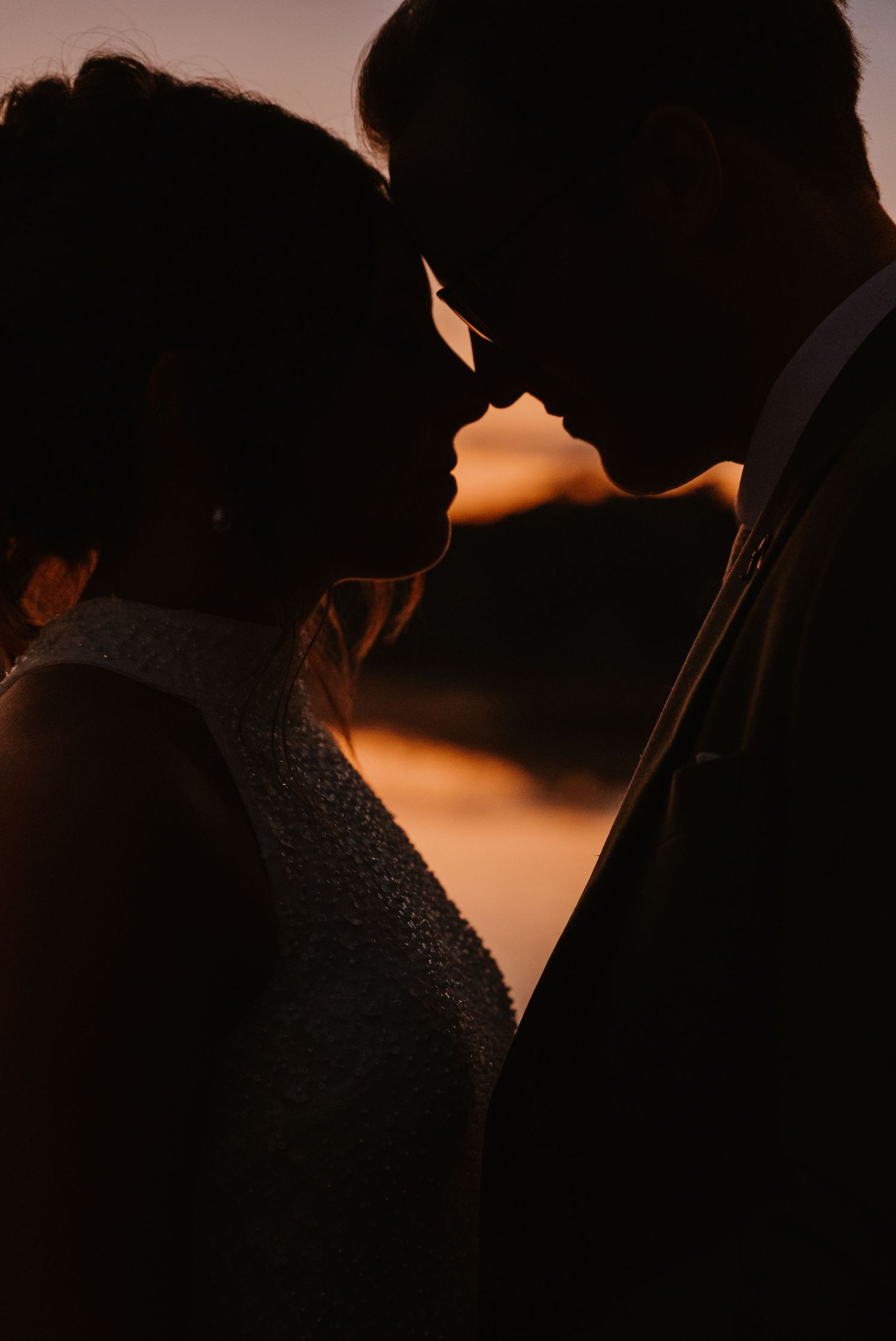 Romantic silhouette of bride and groom touching foreheads against a warm golden sunset sky.