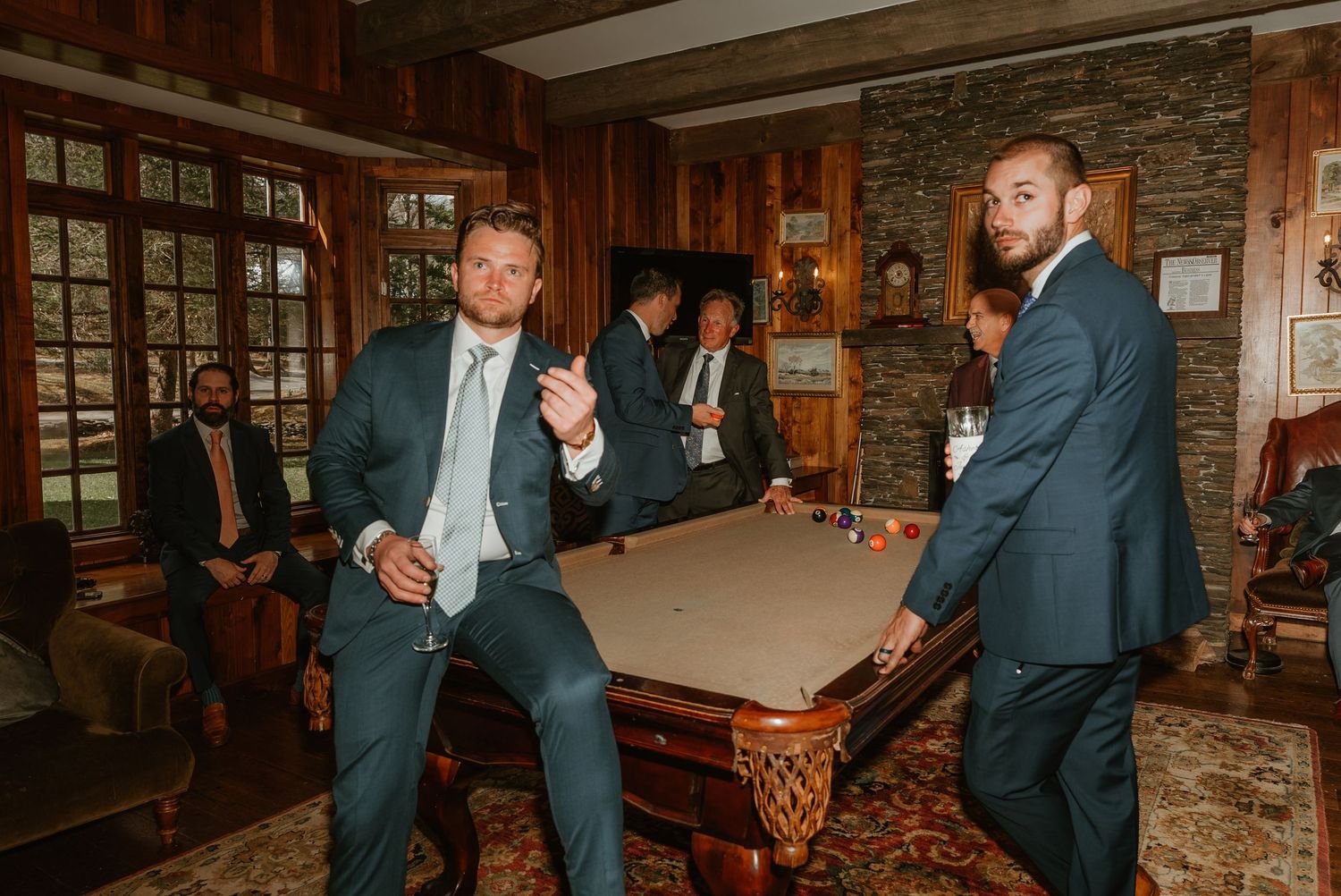 Groomsmen in navy suits socialize around a pool table in a wood-paneled room during wedding reception.