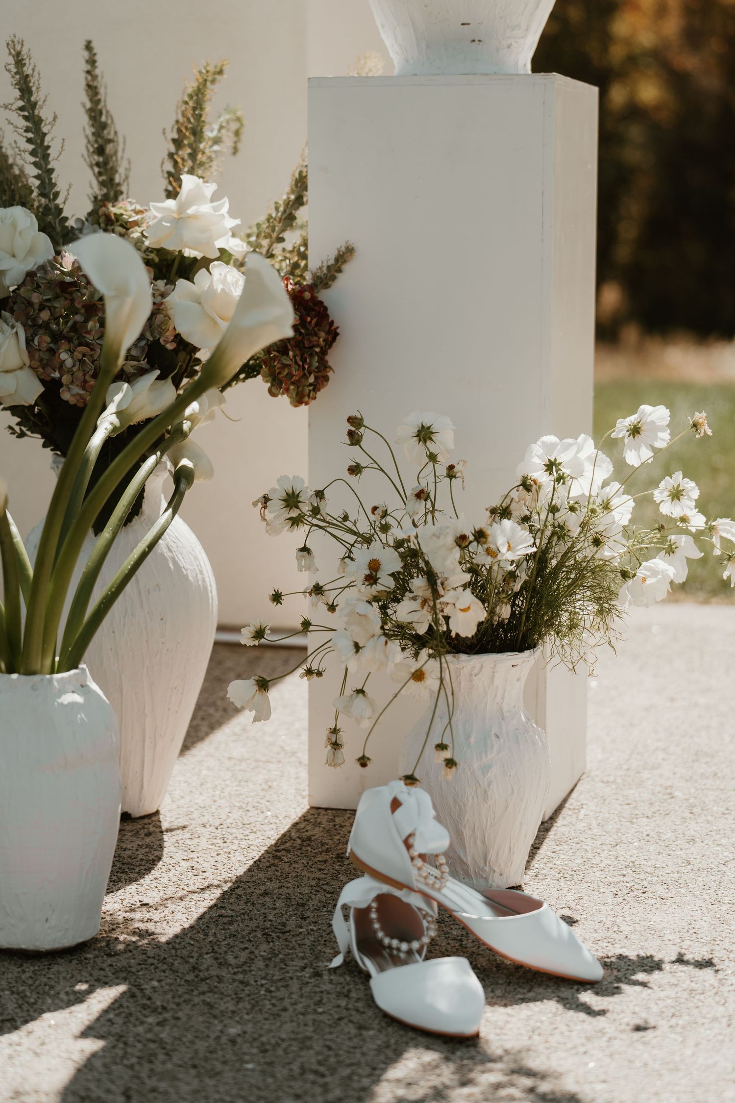 White ceramic vases with wildflowers and bridal heels displayed on sunlit outdoor wedding ceremony decor.