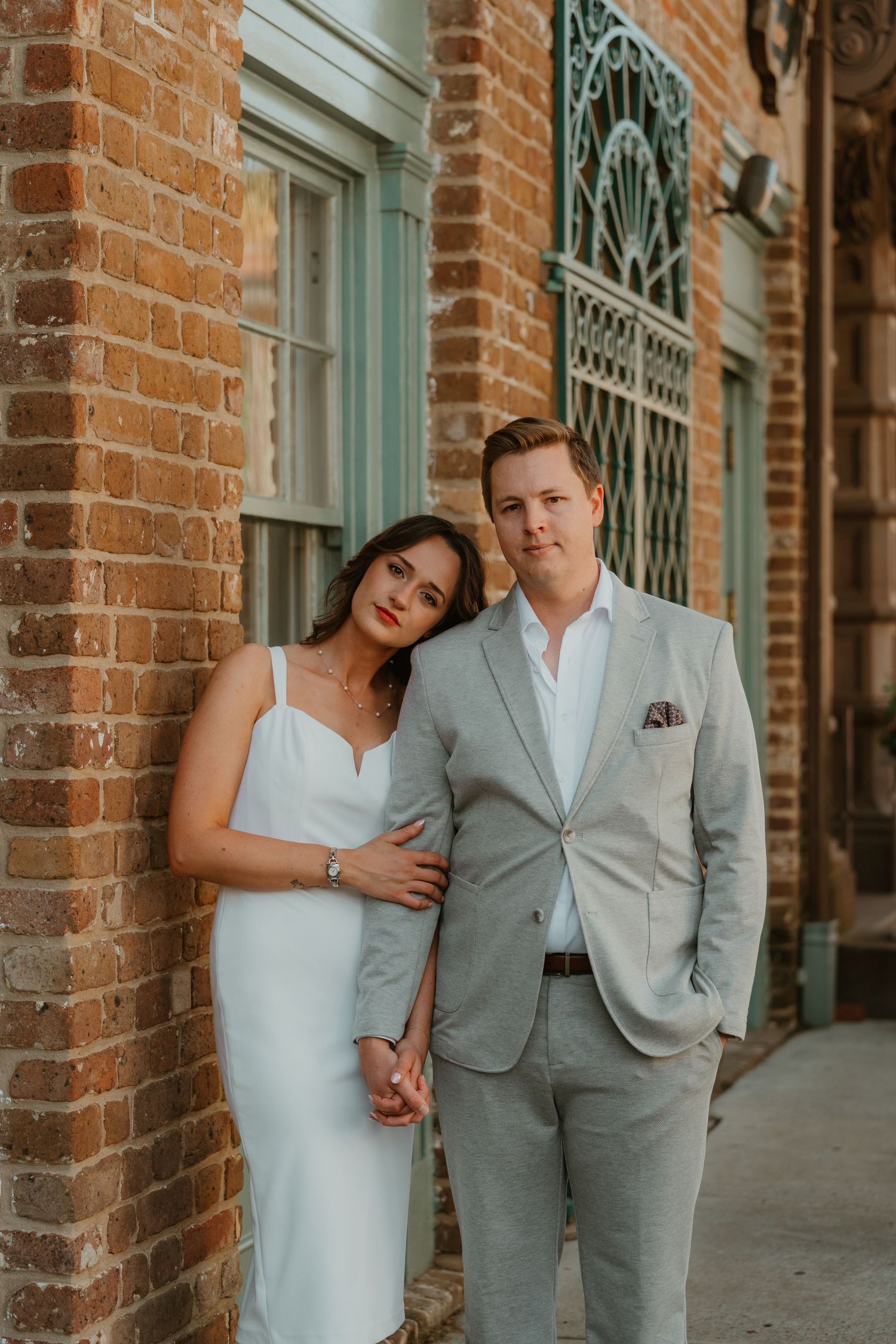 Elegant couple posing against a brick wall, woman in white dress and man in grey suit for wedding portraits.