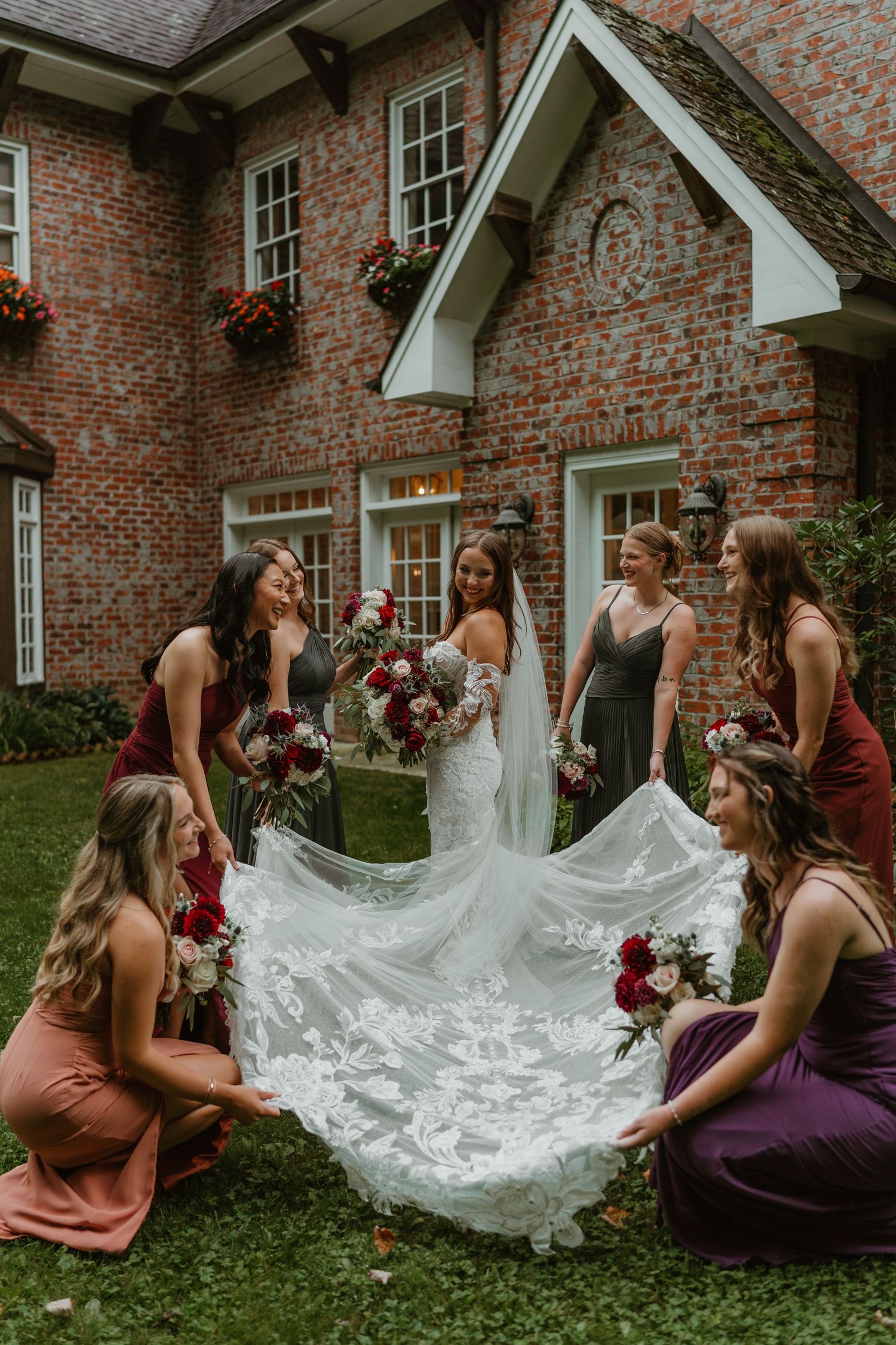 Bride in lace gown surrounded by bridesmaids in jewel-toned dresses holding bouquets outside brick venue.