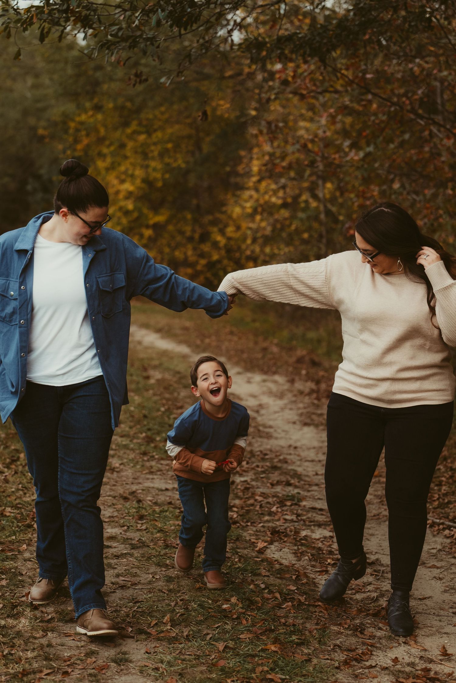 Two moms swing their laughing young son between them on a colorful autumn forest trail.