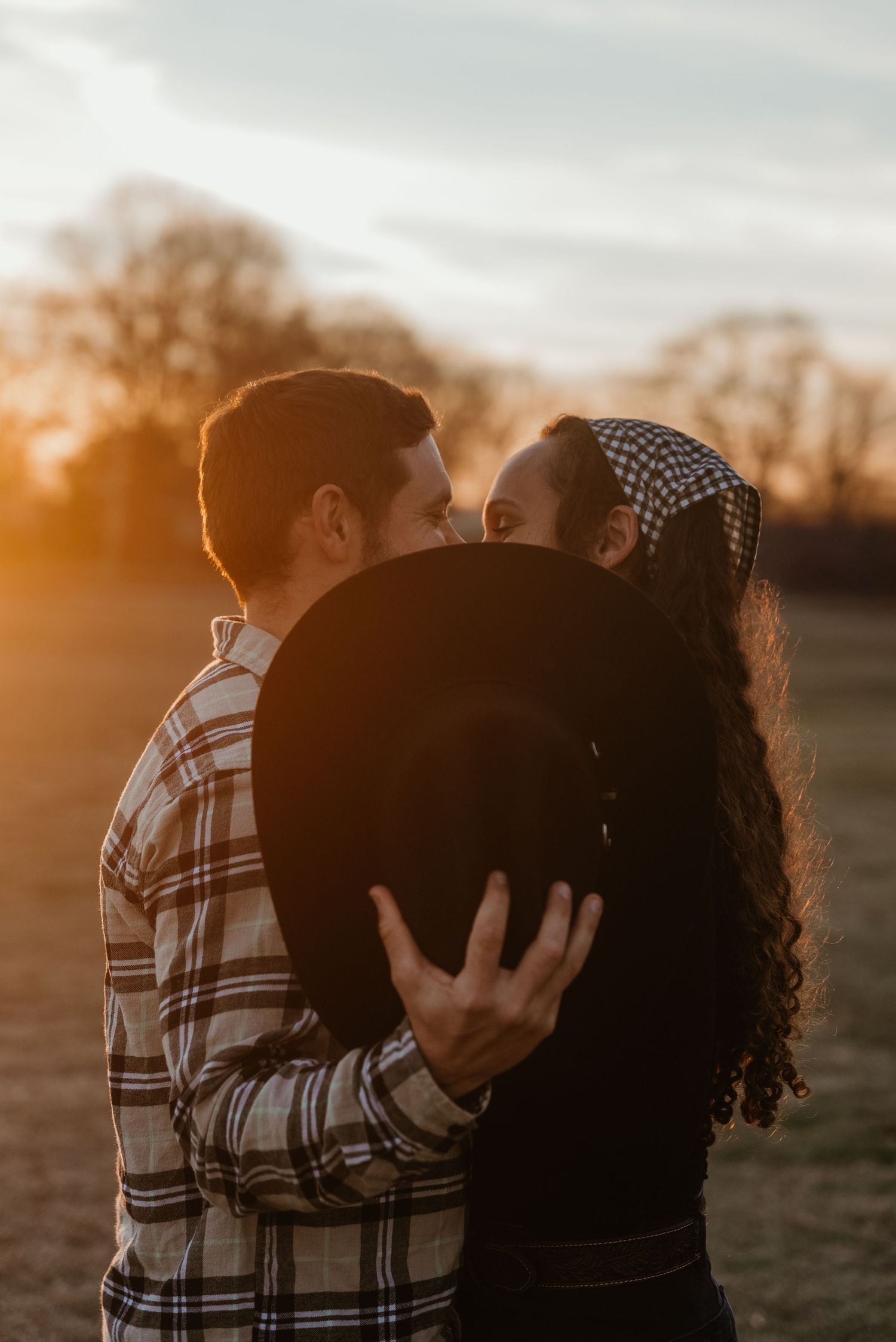 Couple embracing and kissing at sunset in an open field, backlit by warm golden light.