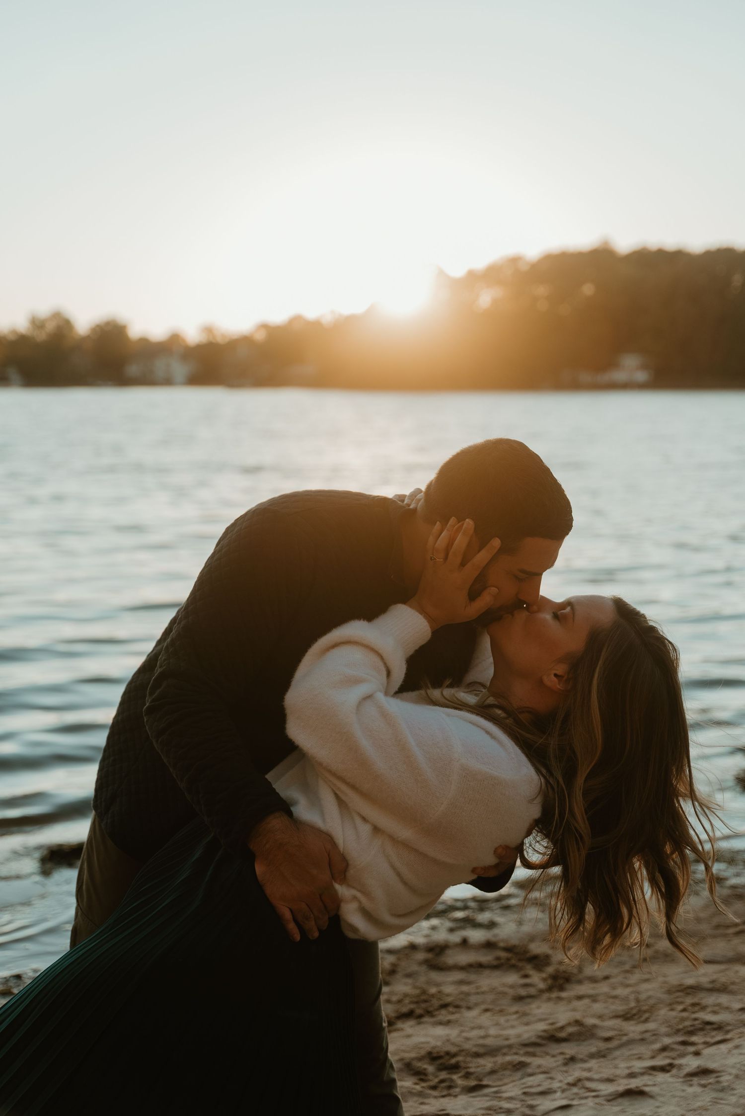 Couple sharing a romantic dip kiss on a sandy beach at golden sunset by the water.