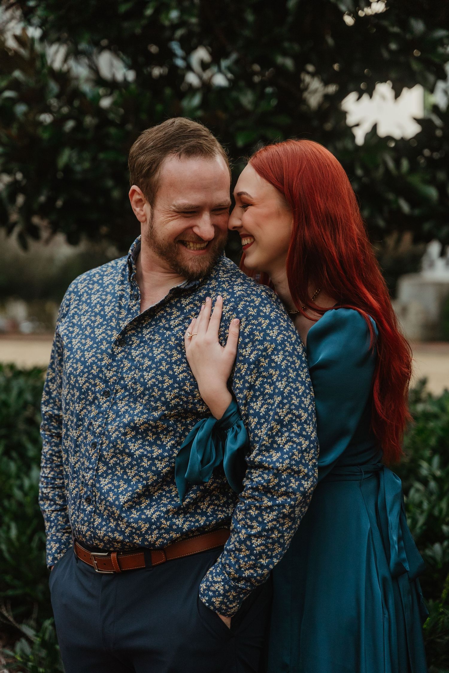 Laughing couple embracing outdoors, man in floral shirt and woman in teal dress in a garden.