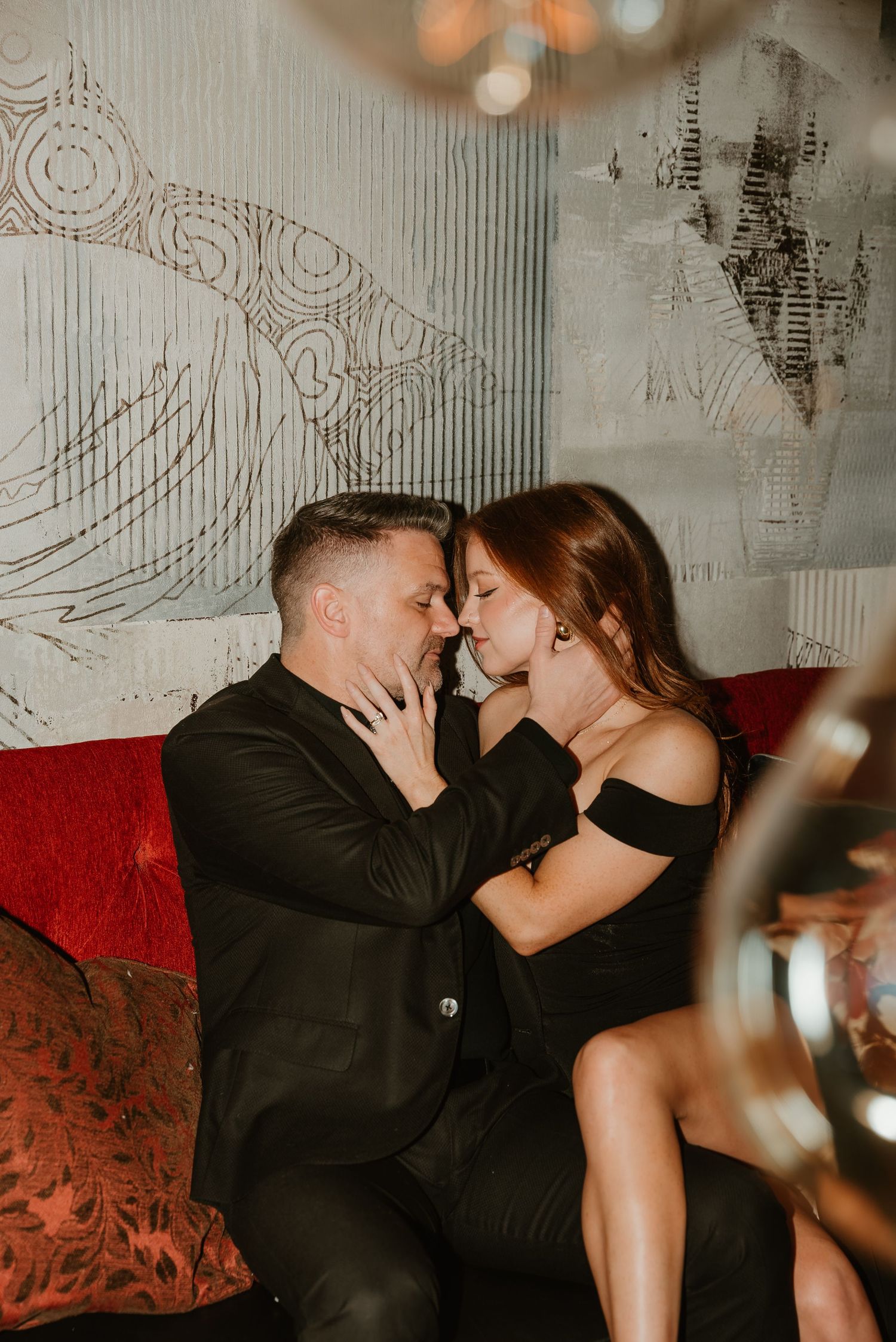 Couple leaning in for a kiss on a red velvet couch in an elegant dark lounge.