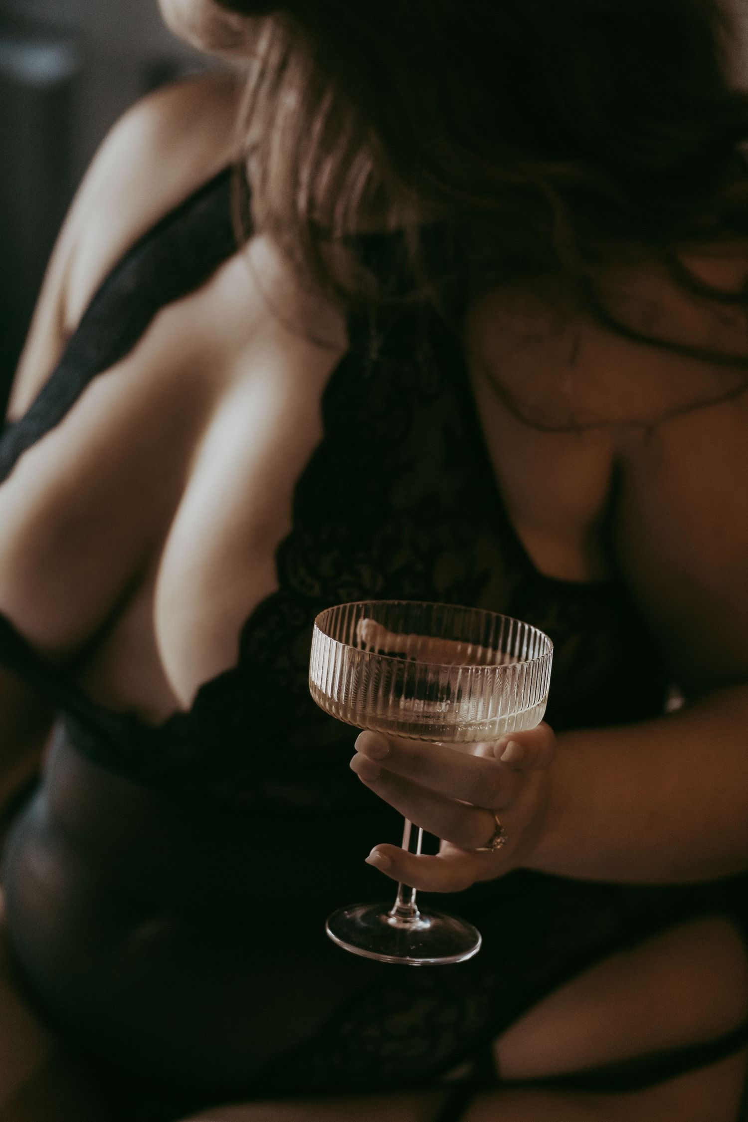 A woman in black lace lingerie holds an elegant coupe glass in a moody, intimate boudoir setting.