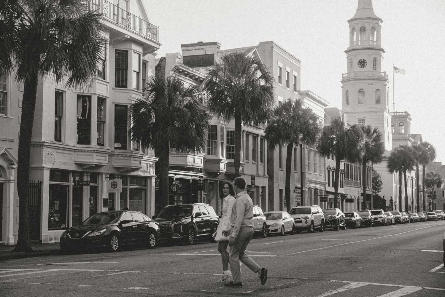 Black and white photo of couple crossing a palm-lined street near historic church.