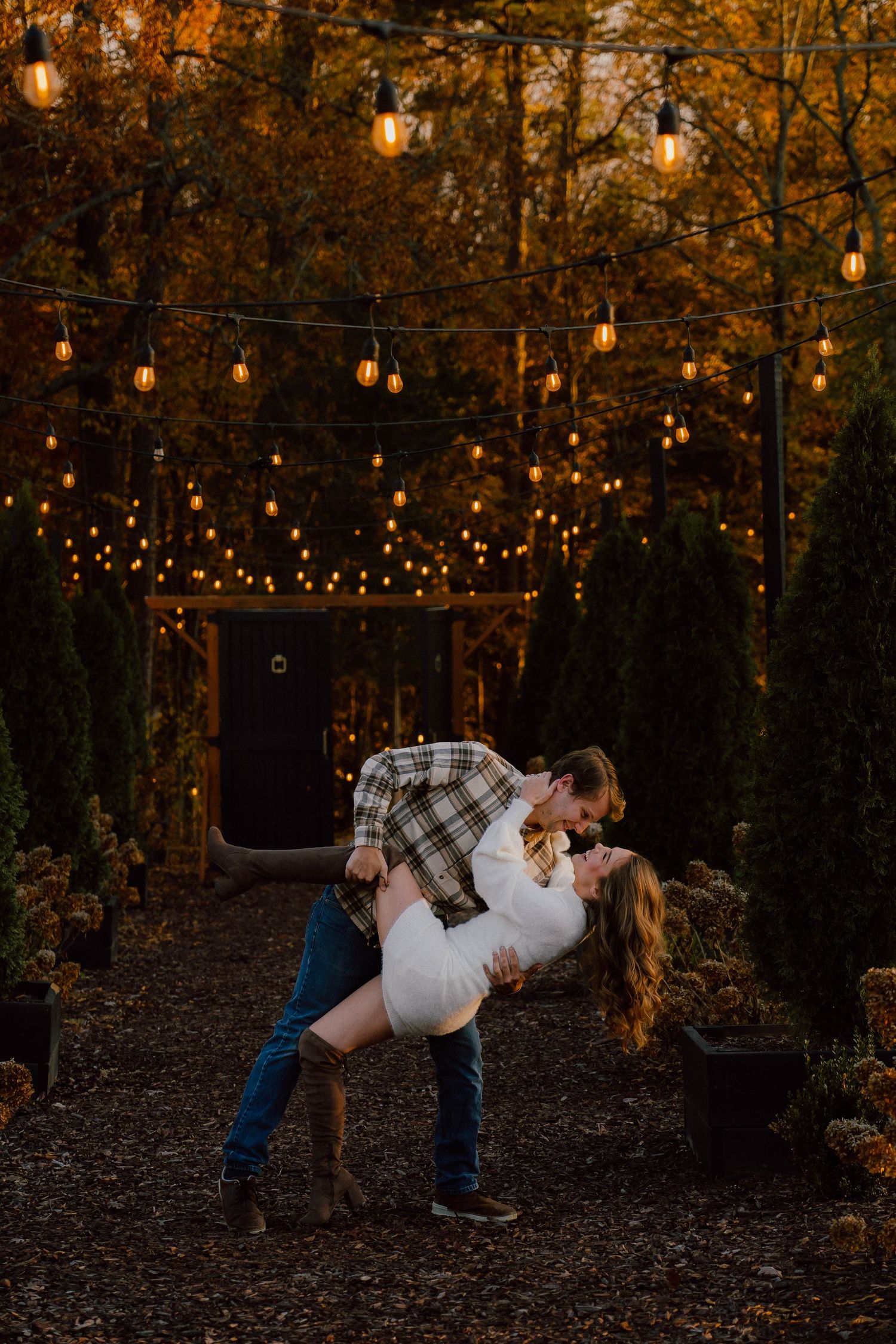Couple sharing a romantic dip kiss under string lights in an autumn outdoor setting at night.