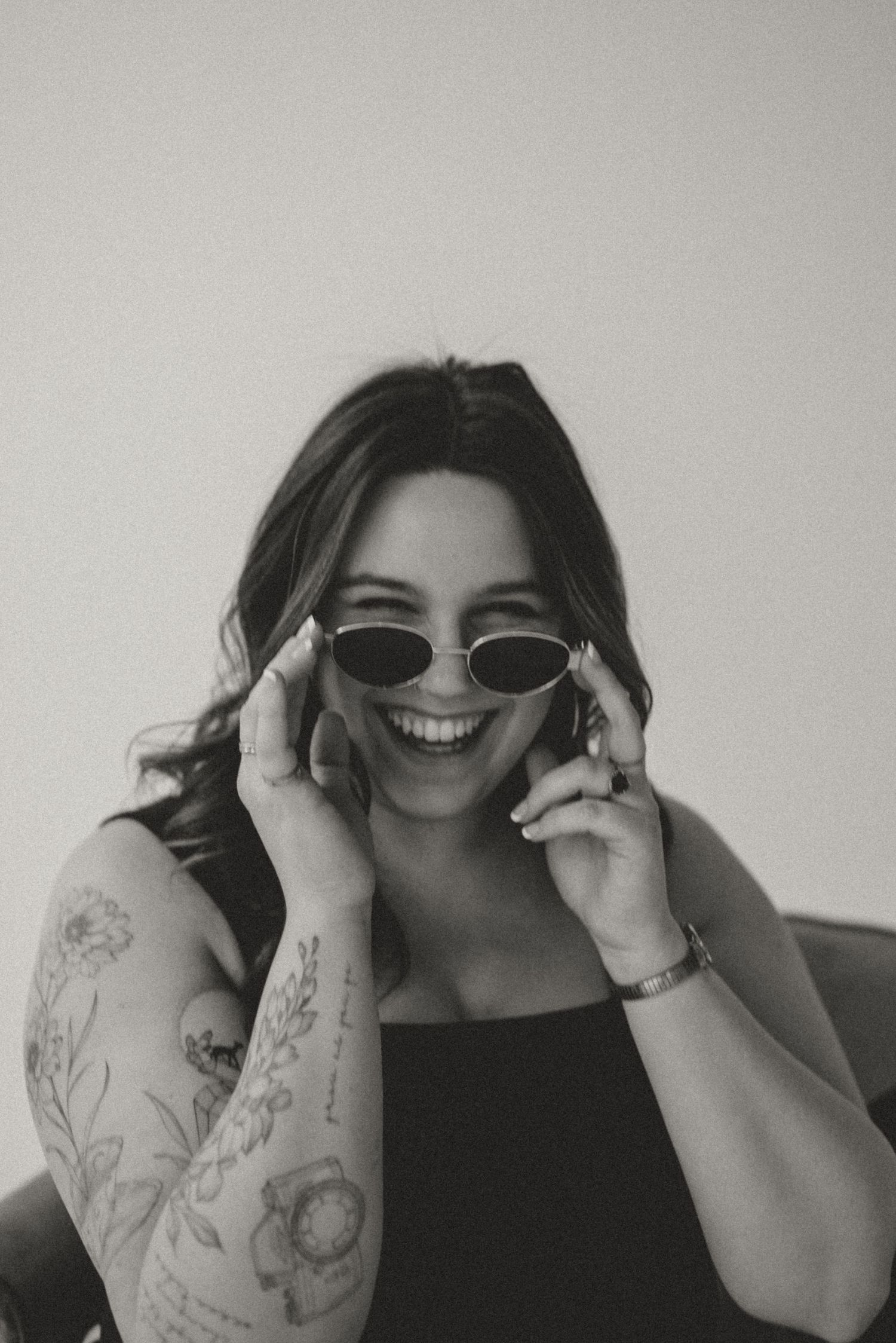 Smiling woman in black top with floral arm tattoo posing with oval sunglasses in black and white.